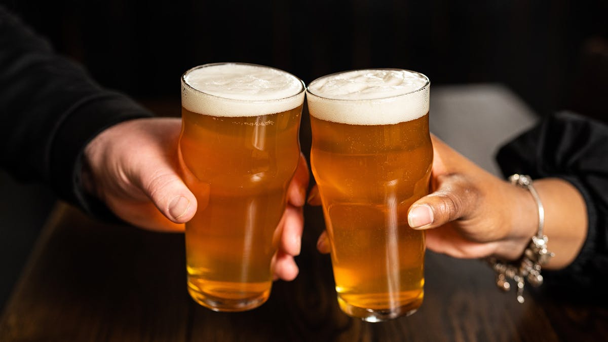 two people holding a pint of beer for National Beer Day at Rivercrest in Astoria, Queens with their one time only deal: buy one draft beer, get the second for 5 cents only on Friday, April 7th, 2023