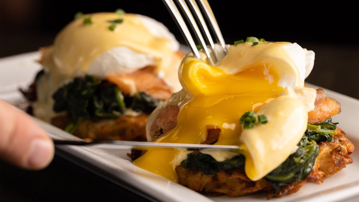 cutting into the poached egg of our Eggs Benedict Florentine brunch dish at Astoria's Rivercrest