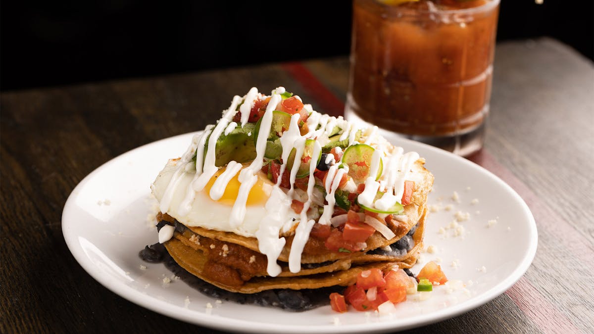 a plate of Huevos Rancheros on a table with a blood mary in the back for bottomless brunch