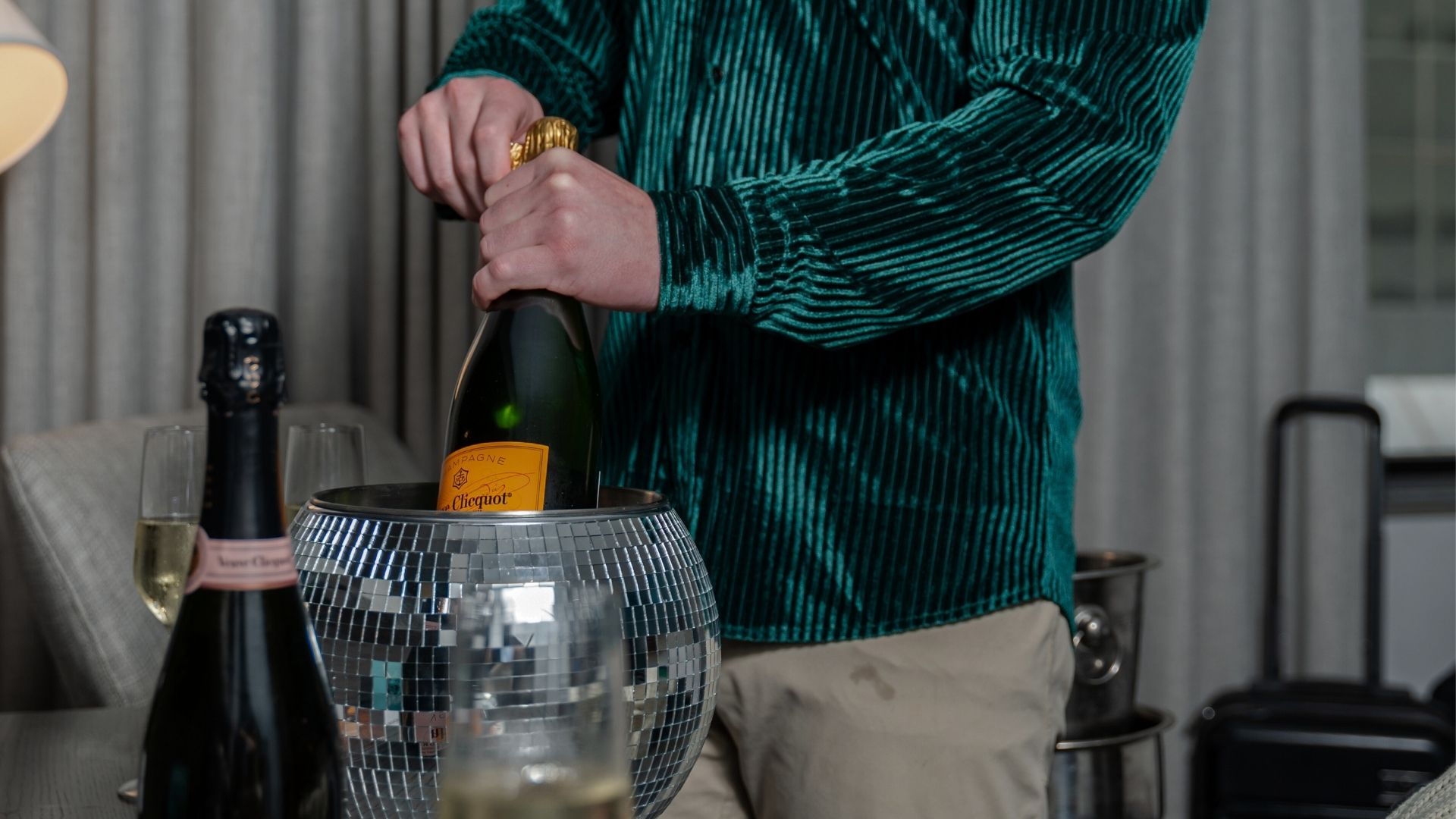Guest opening a bottle of Veuve Clicquot champagne over a mirrored ice bucket in a hotel room, with flutes and luggage visible in the background.