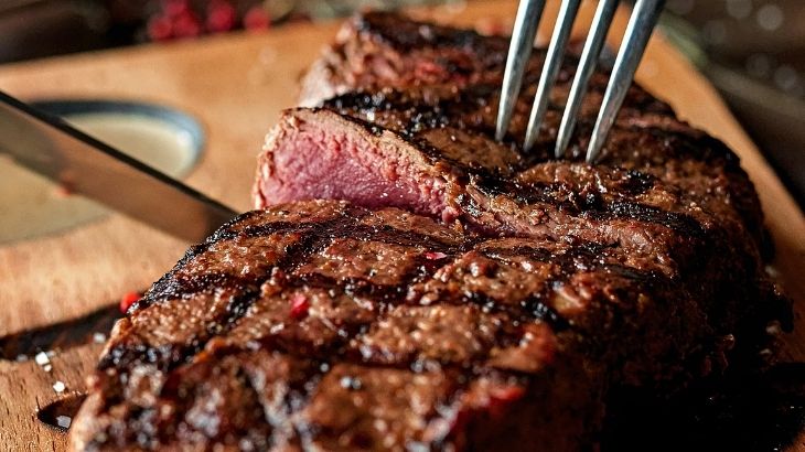 Close-up of a grilled steak sliced on a wooden board, showing a juicy medium-rare center with a fork and knife cutting into it.