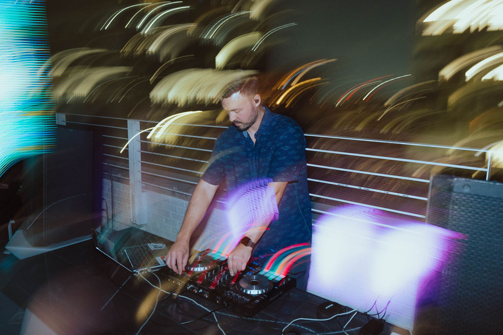 DJ performing at a rooftop event at night, mixing music on a controller with colorful light trails and city skyline in the background.