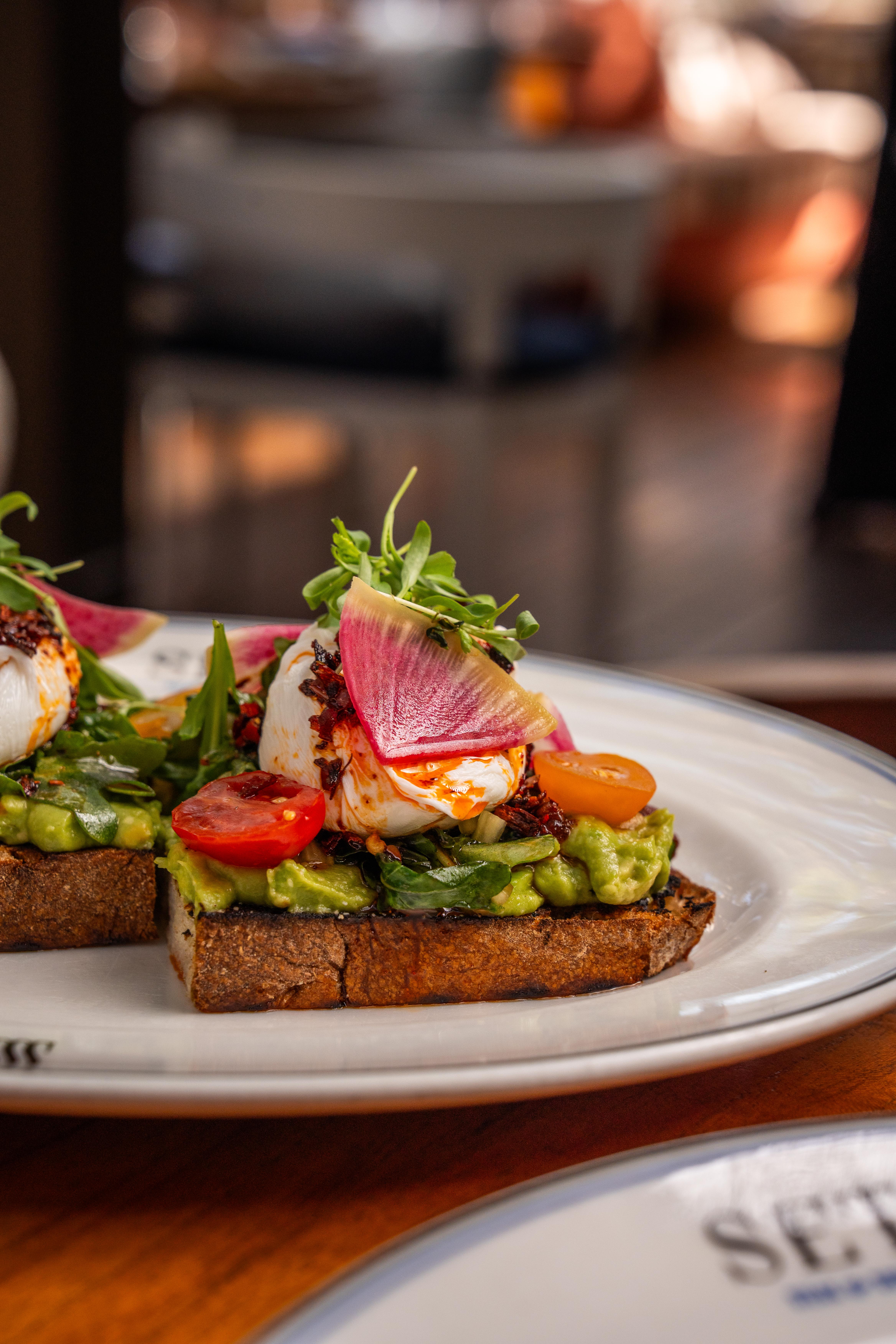 Avocado toast with tomatoes, radish sitting on a white plate on a wooden table.