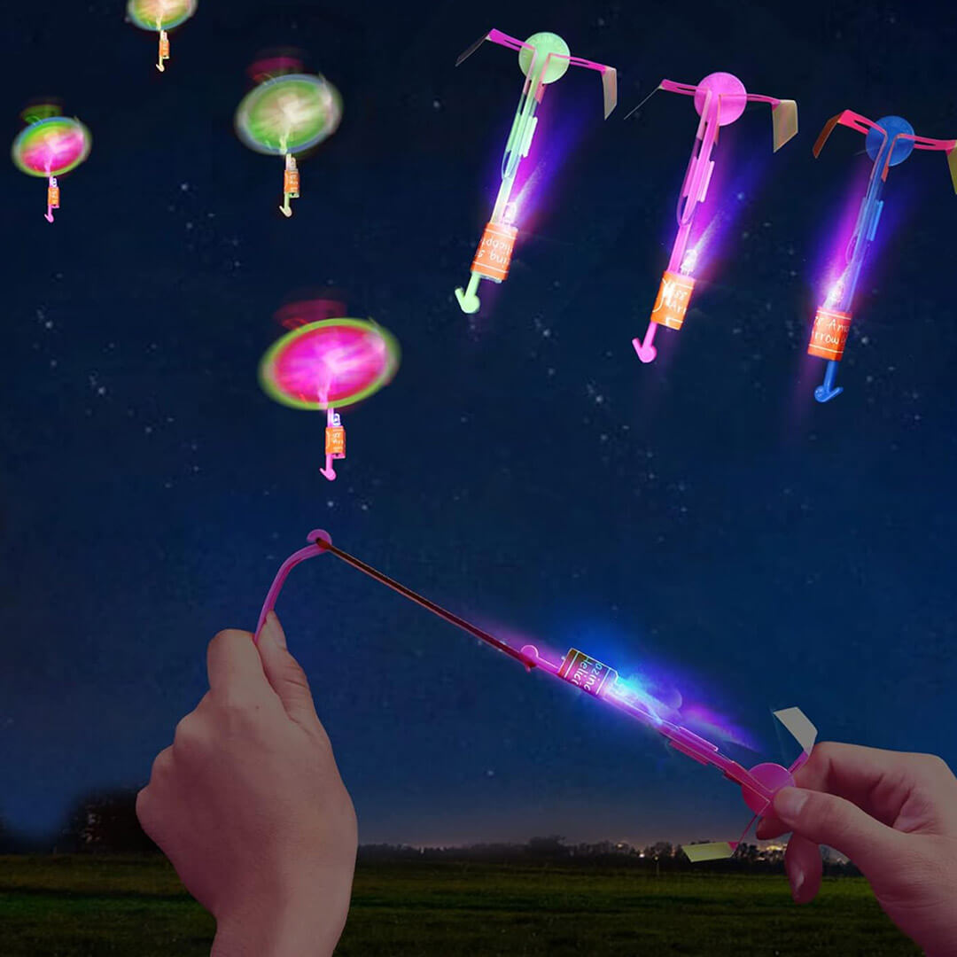 Hands using a slingshot band to launch glowing LED arrow helicopter toys into a dark blue night sky.
