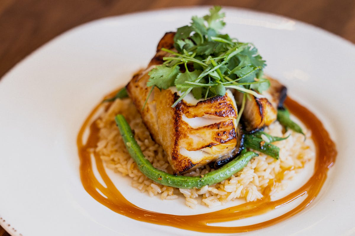 Misoyaki Glazed Cod with green beans, coconut rice and eel sauce with green onion and daikon sprouts with wine bottles and salad in background.