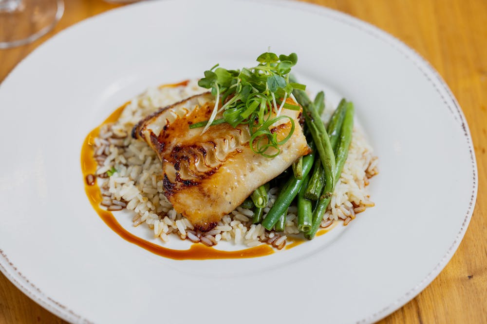 Misoyaki Glazed Cod with green beans, coconut rice and eel sauce with green onion and daikon sprouts with wine bottles and salad in background.