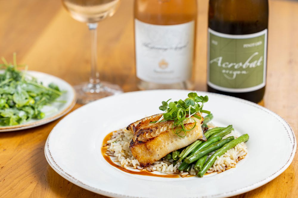 Misoyaki Glazed Cod with green beans, coconut rice and eel sauce with green onion and daikon sprouts with wine bottles and salad in background.