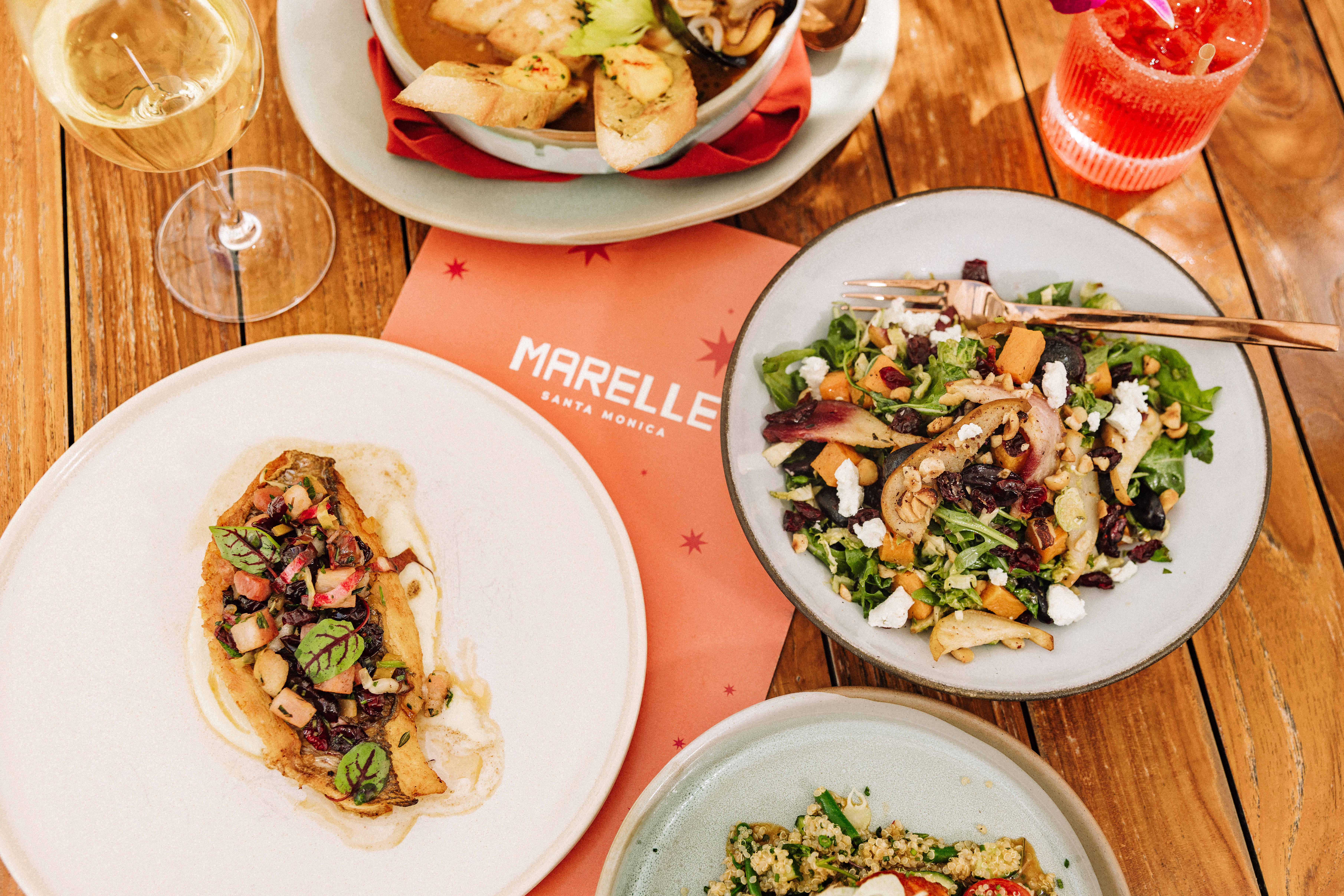 A rustic wooden table set with Marelle Santa Monica’s fall menu dishes, including a fresh autumn salad with pears, squash, and goat cheese, a golden seared fish topped with seasonal vegetables, a hearty soup with toasted bread, and a glass of white wine alongside a vibrant red cocktail, with a Marelle-branded menu in the center