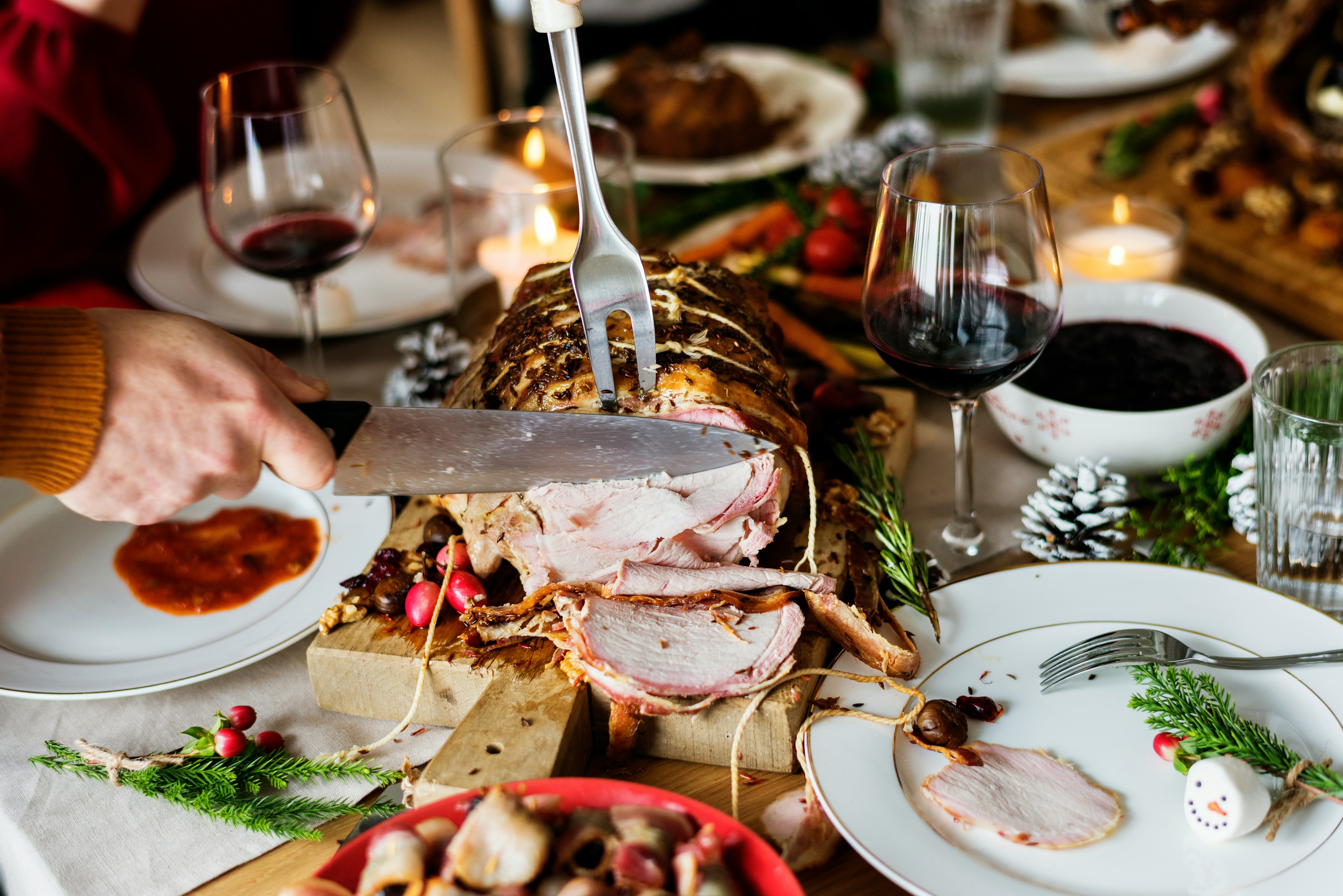 A person carving a roasted meat dish at a festive dinner table, surrounded by glasses of red wine, plates, candles, greenery, pinecones, and holiday decorations.
