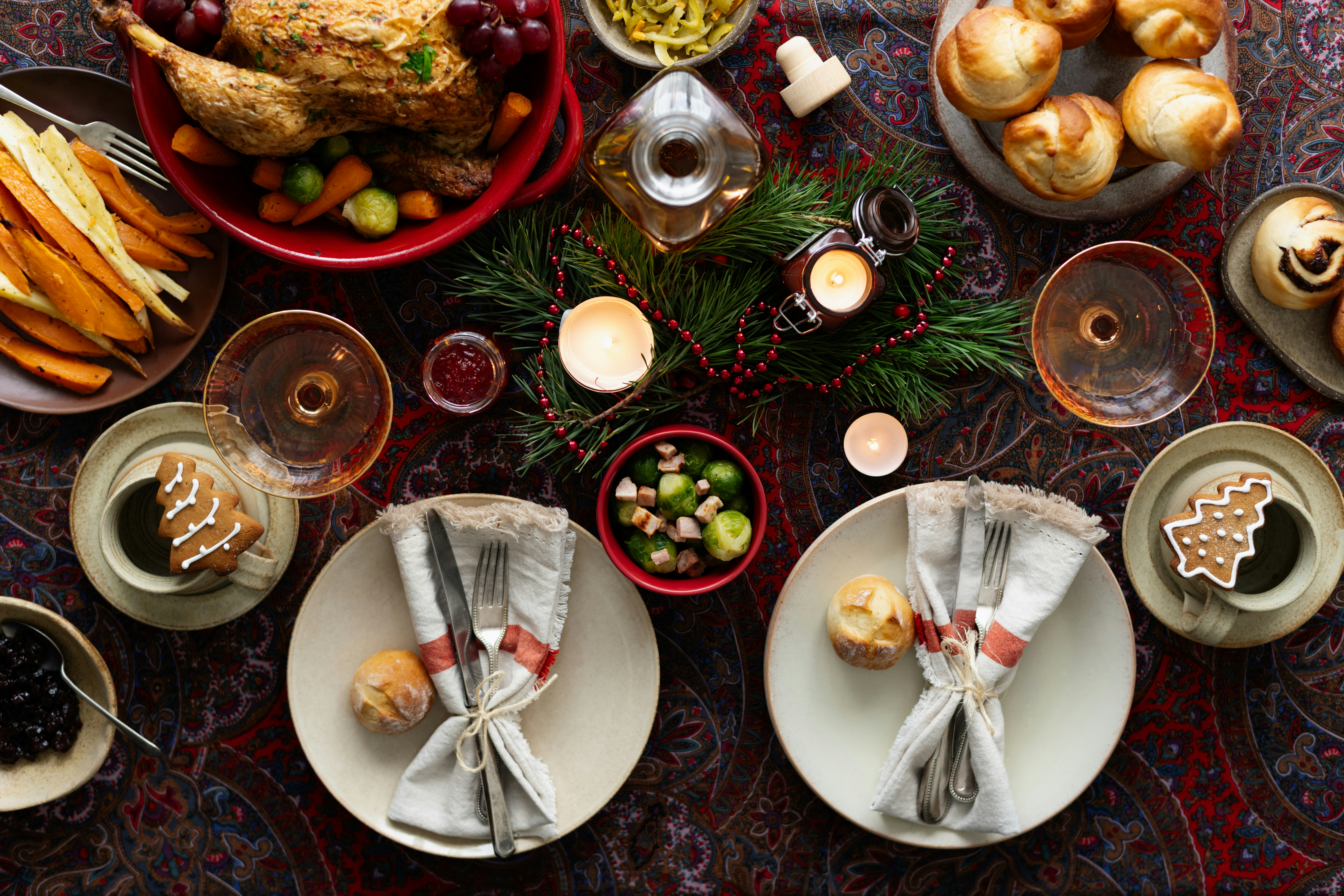 An overhead view of holiday themed dinner table featuring whole turkey, dinner plates with wrapped silverware, greenery and red beads
