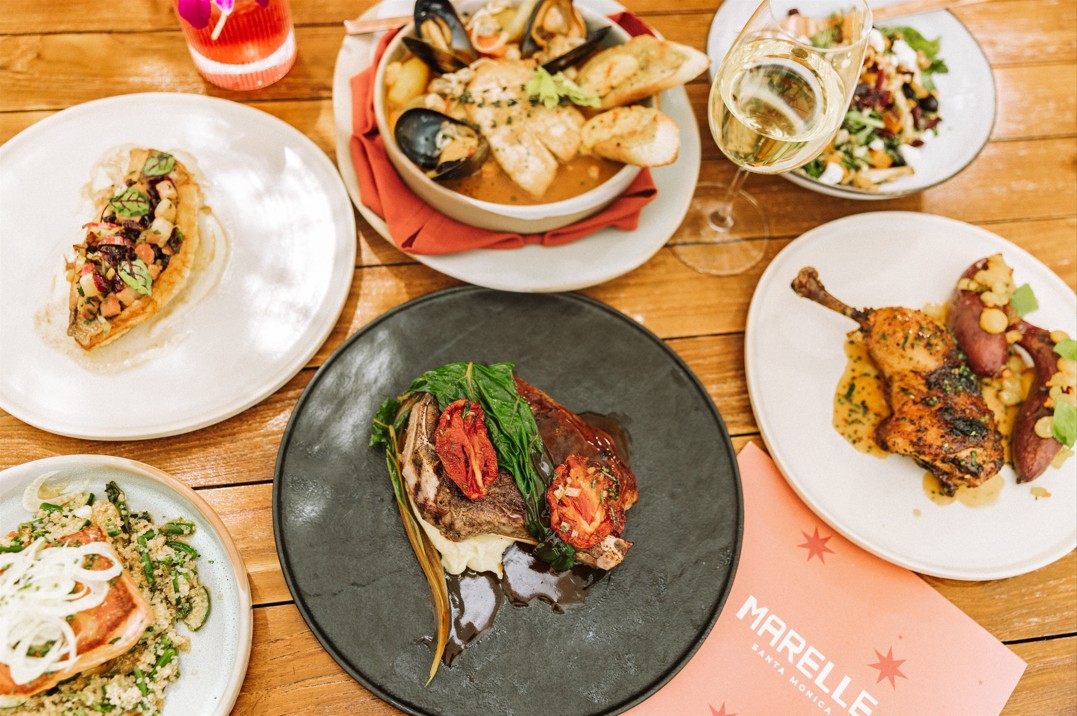 An overhead view of Marelle Santa Monica’s fall menu spread on a wooden table, featuring a seared fish topped with seasonal vegetables, a bowl of seafood stew with toasted bread, a grilled chicken dish with fingerling potatoes, a hearty steak with greens and roasted tomatoes, a salmon over couscous, a fresh autumn salad, and glasses of white wine and a red cocktail