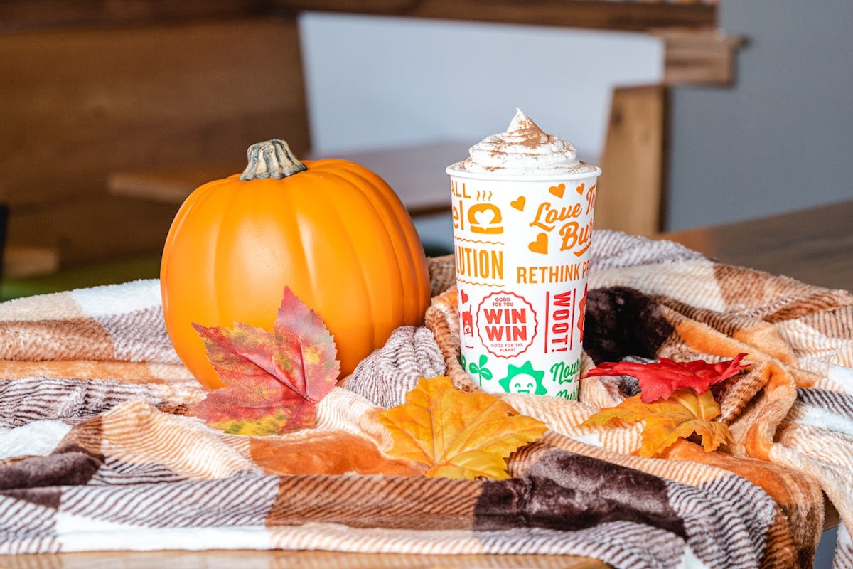 A festive autumn setup featuring a bright orange pumpkin, colorful fall leaves, and a whipped cream–topped pumpkin spice shake in a branded cup, all arranged on a soft plaid blanket with warm brown, orange, and cream tones.