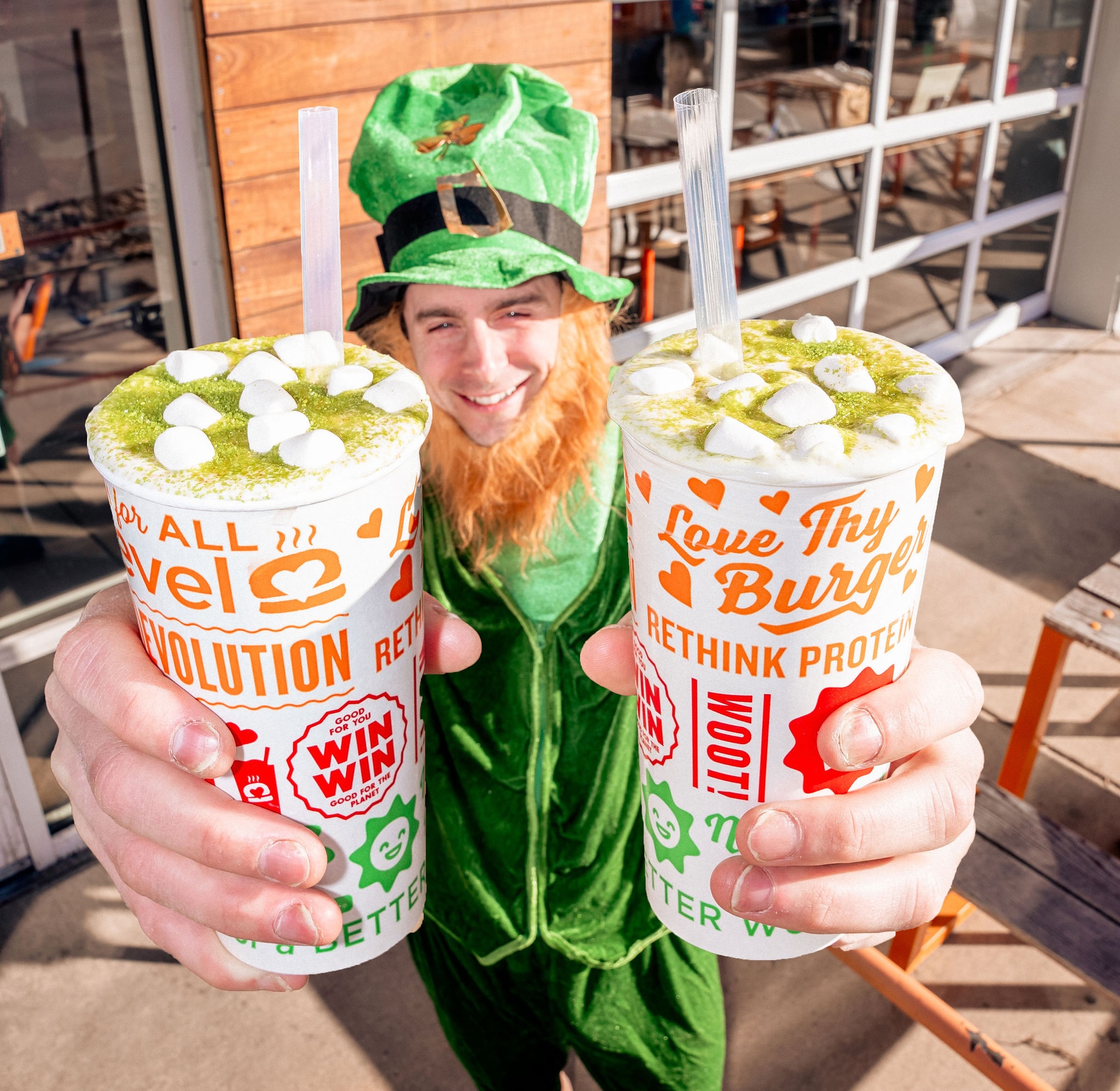 A festive leprechaun holding two Lucky Leprechaun Shakes from Next Level Burger, topped with mini marshmallows