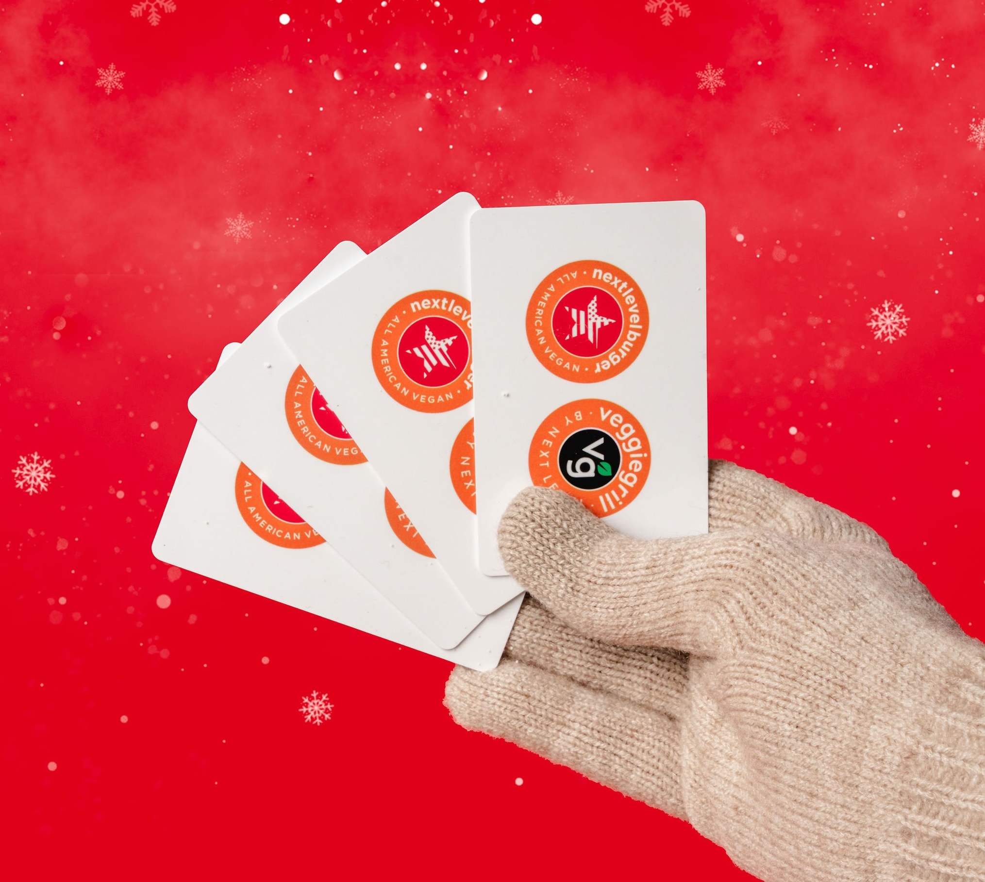Gloved hand holding Next Level Veggie Grill gift cards against a red holiday background with snowflakes.