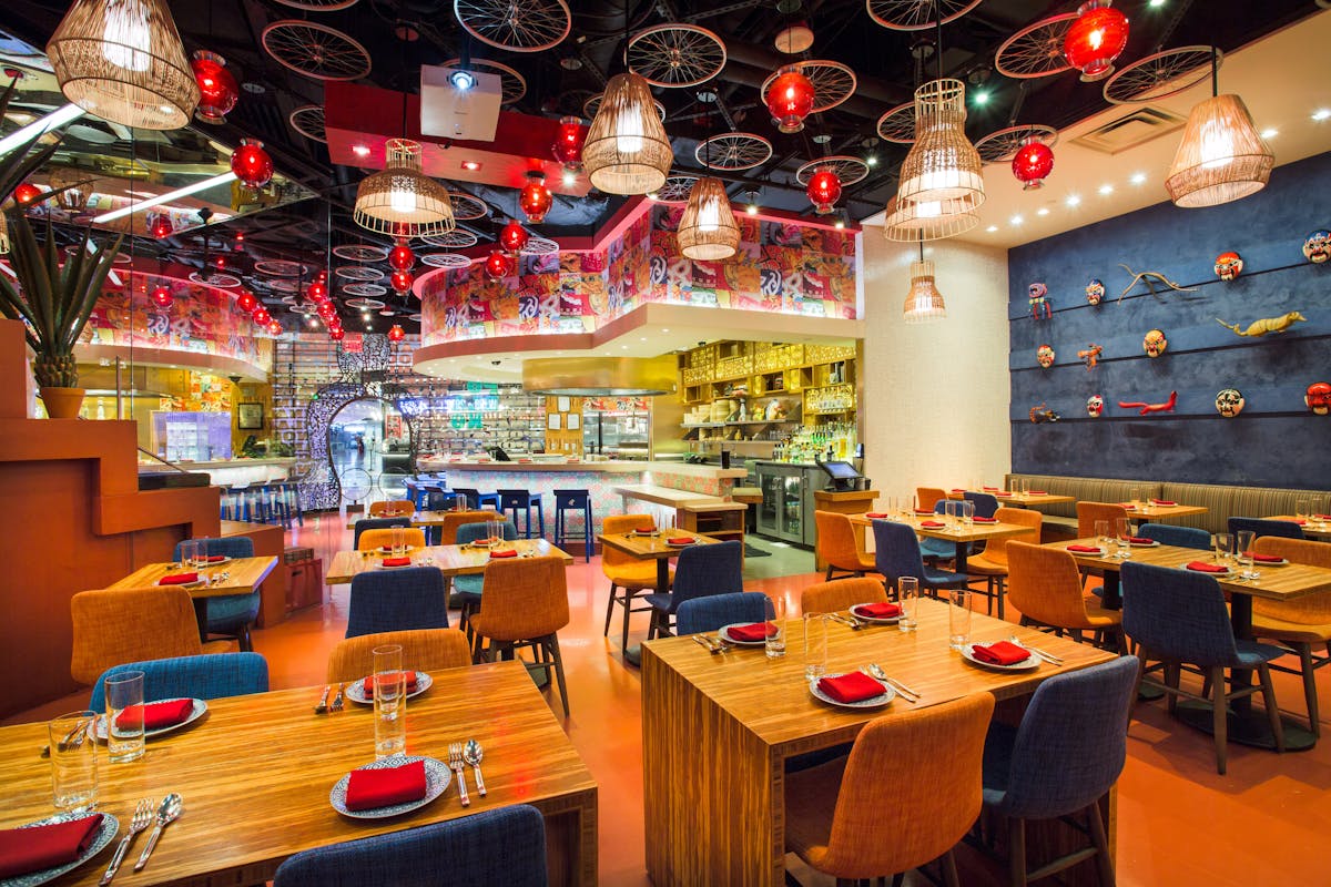 a dining room with table seating, blue and yellow chairs, red tablecloths, and red lighting accents on the cieling.
