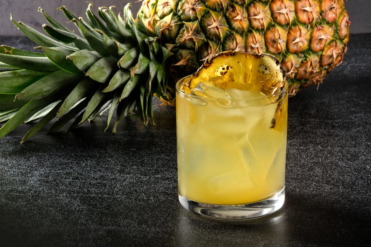 a cocktail with a wedge of pineapple, with a whole pineapple laying behind the cocktail.