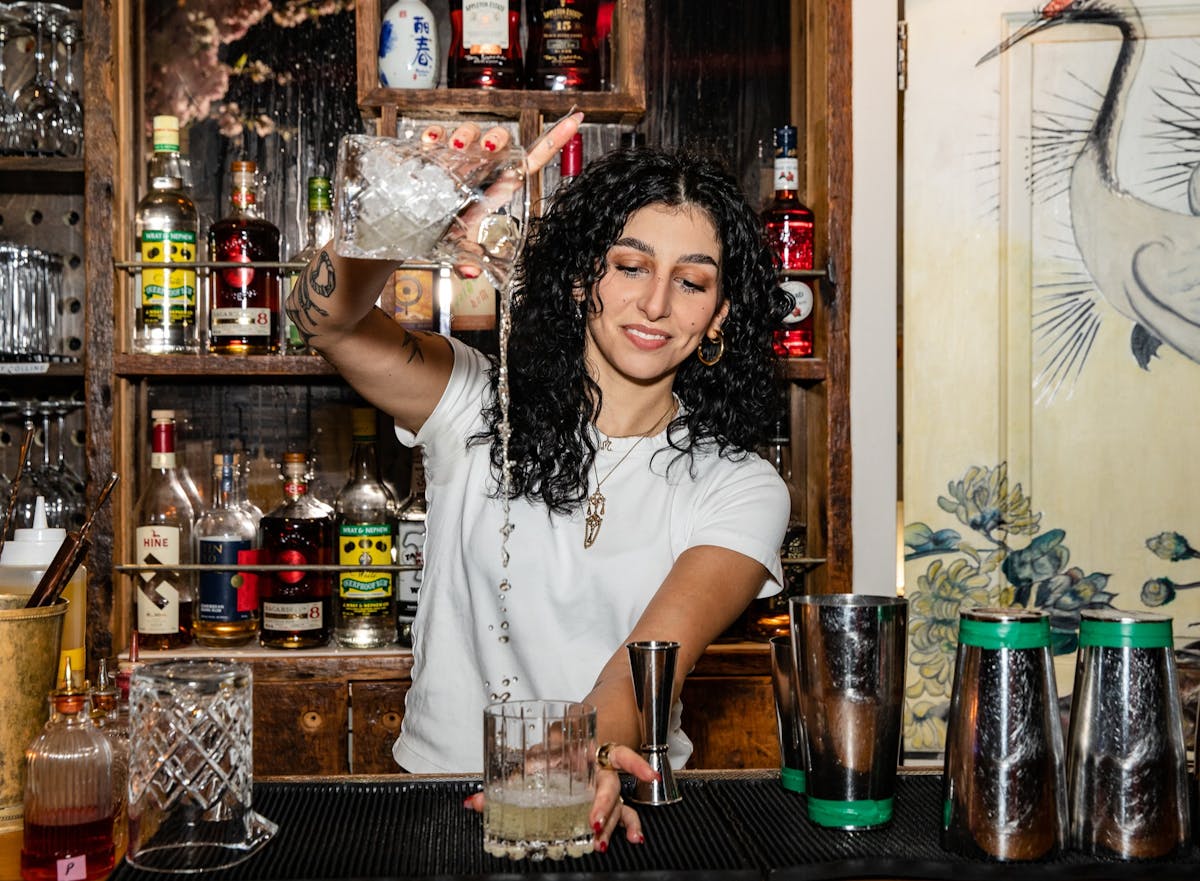 a woman with long curly hair smiling while making a cocktail