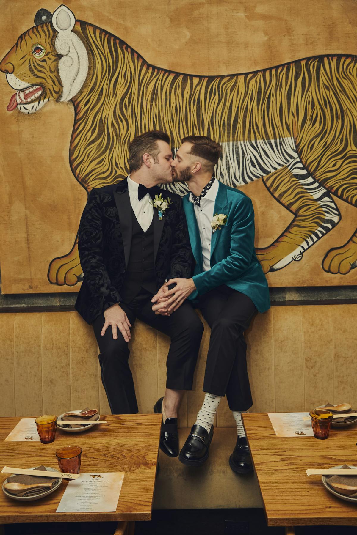 a groom wearing a black blazer and a groom wearing a teal blazer kissing on top of a booth