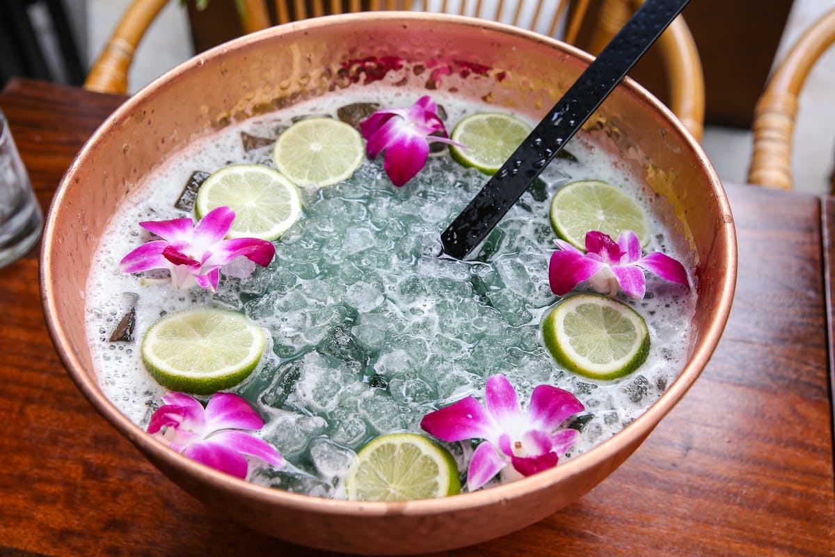 a large format cocktail in a copper bowl with ice, flowers, and lime