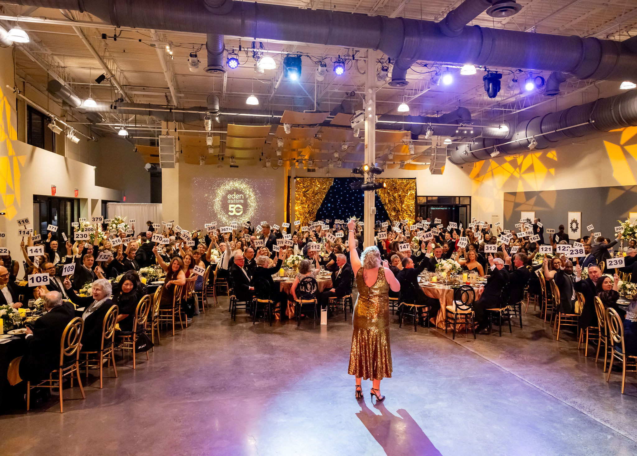 a woman in a gold dress standing on a stage in a large room with tables and chairs
