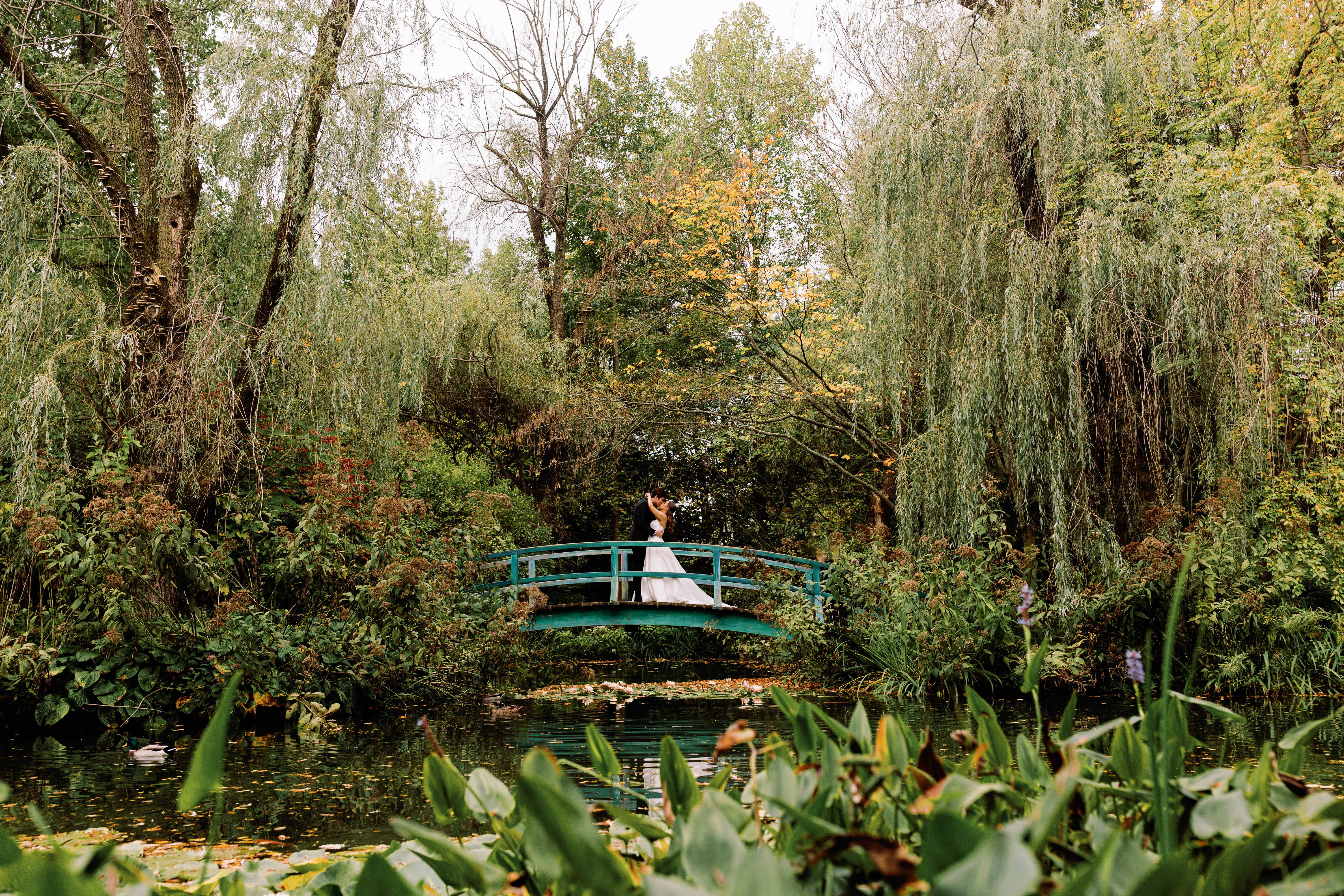 a bride and groom on a bridge over a pond