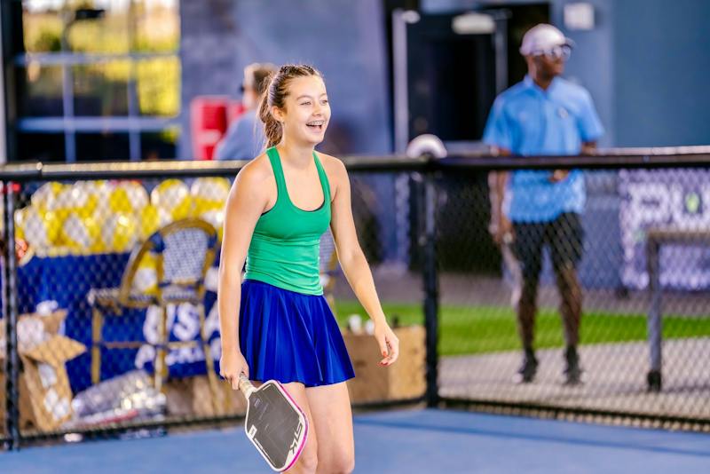 Smiling player in a green top walking onto the court with a paddle at Dink & Dine Pickle Park