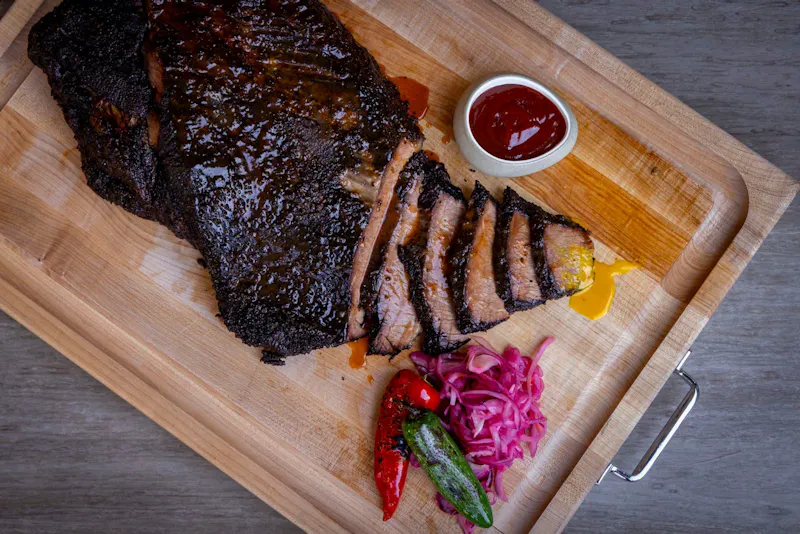 Sliced smoked brisket on a cutting board with pickled onions and BBQ sauce at Dink & Dine Pickle Park