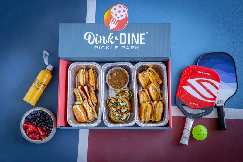 First Smash Breakfast catering box with breakfast sandwiches beside a pickleball paddle at Dink & Dine Pickle Park