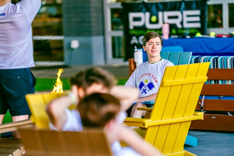 Guests lounging in Adirondack chairs by a fire pit next to the courts at Dink & Dine Pickle Park