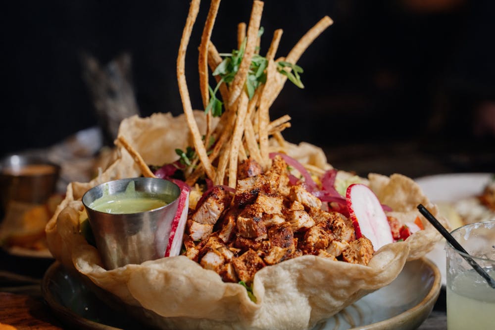 Taco salad in a crispy tortilla bowl filled with grilled chicken, tortilla strips, pickled onions, radish slices, and a side of green avocado sauce at Mijo.