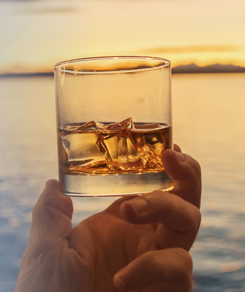 Person's hand holding a bourbon drink with sunset, water views