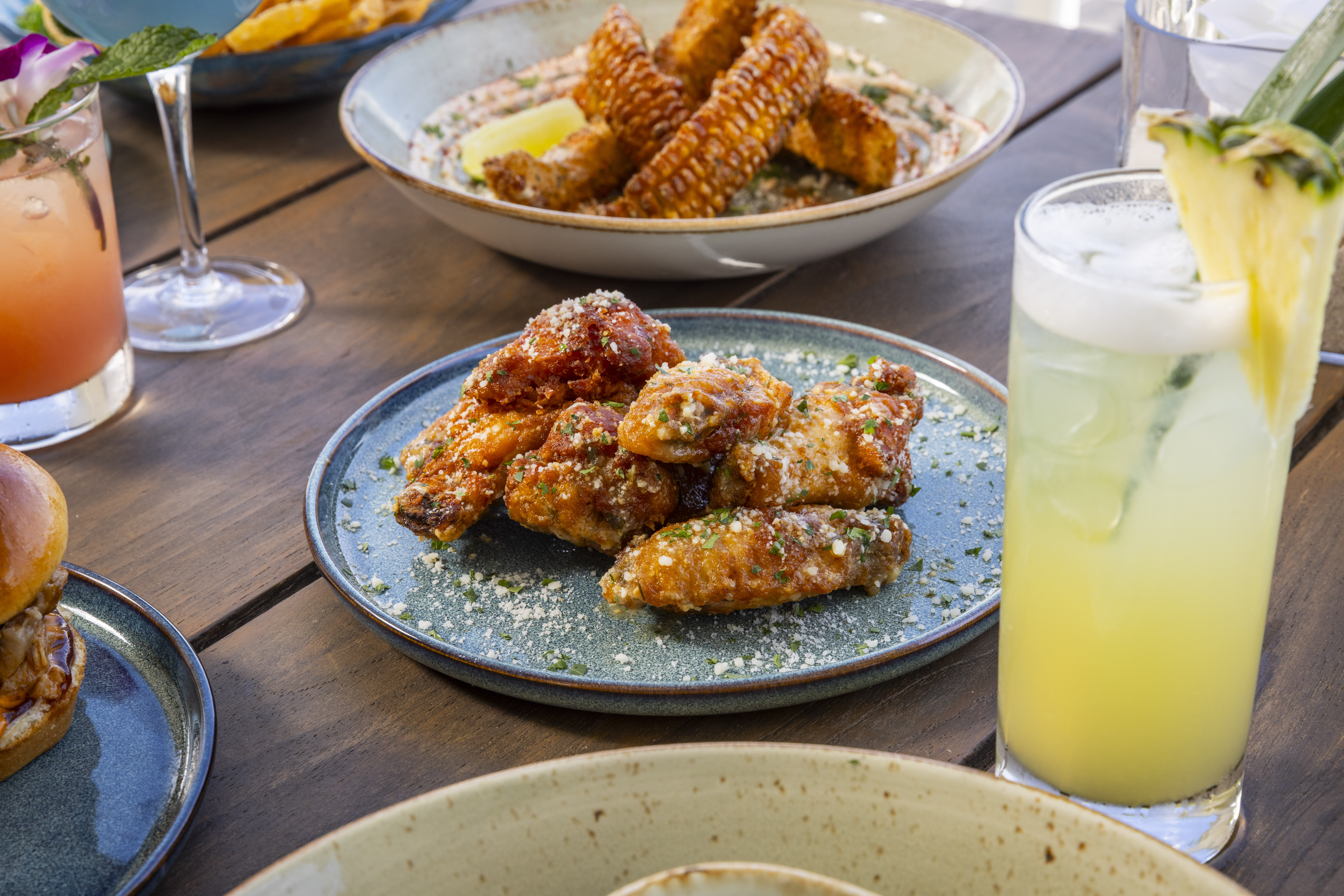 Plate of crispy chicken wings topped with herbs and cheese, paired with tropical cocktails on a wooden table.