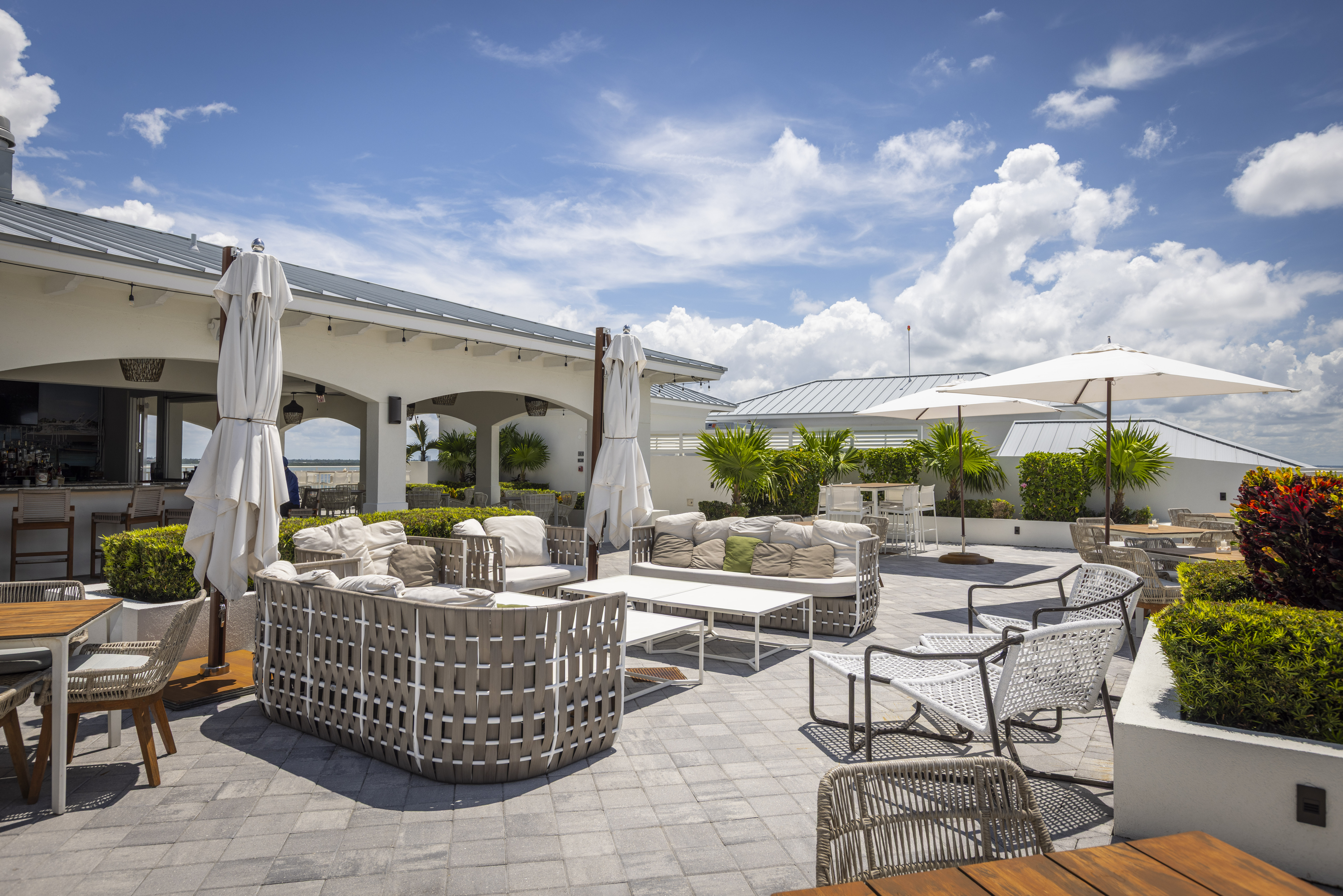 Rooftop lounge with wicker seating, umbrellas, and lush greenery under a bright blue sky.