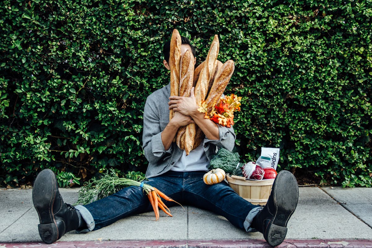 a person sitting with baguettes in front of their face and a basket of produce