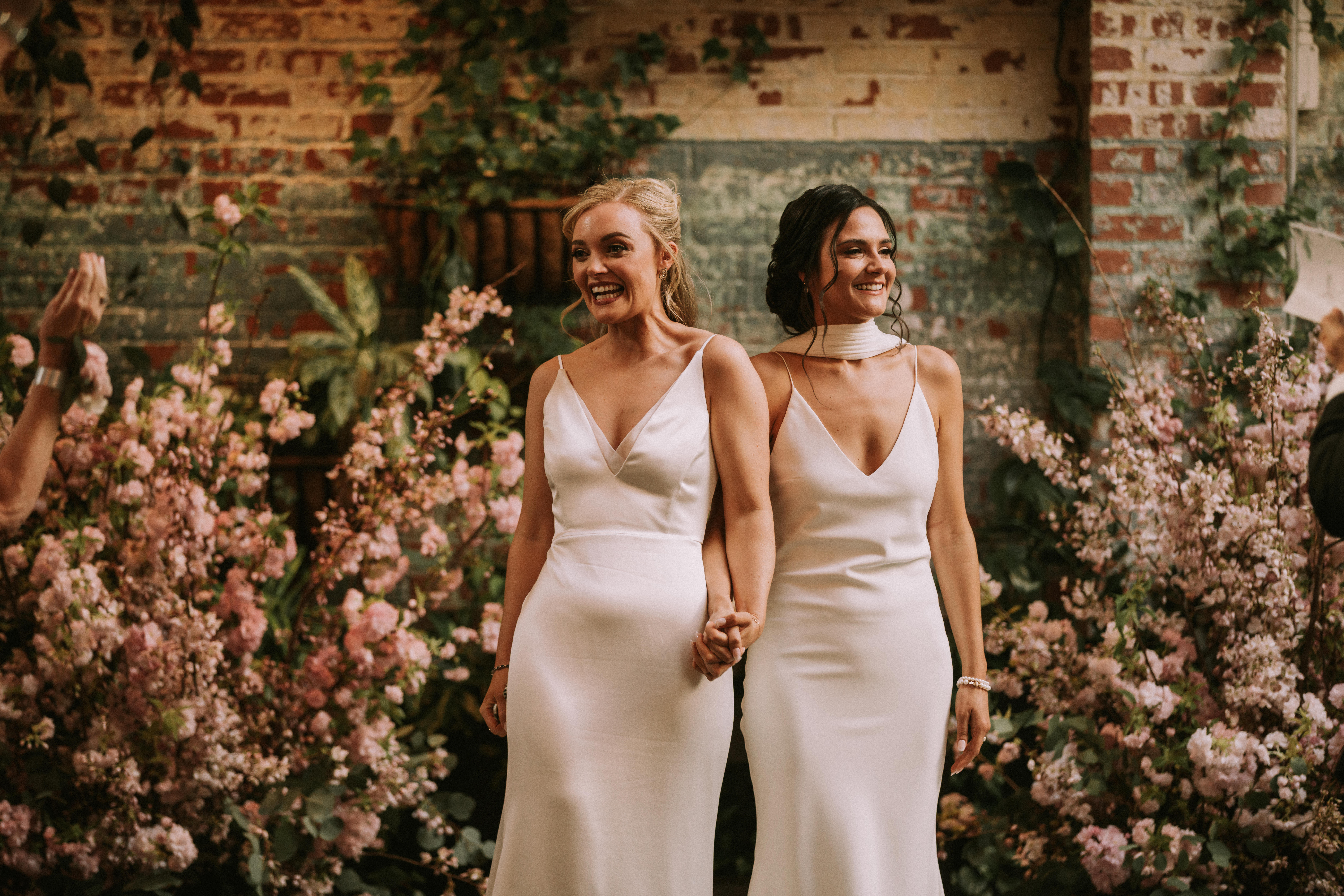 Two newlyed women walk back down the aisle flanked by the beautiful greenery and florals of our living plant wall