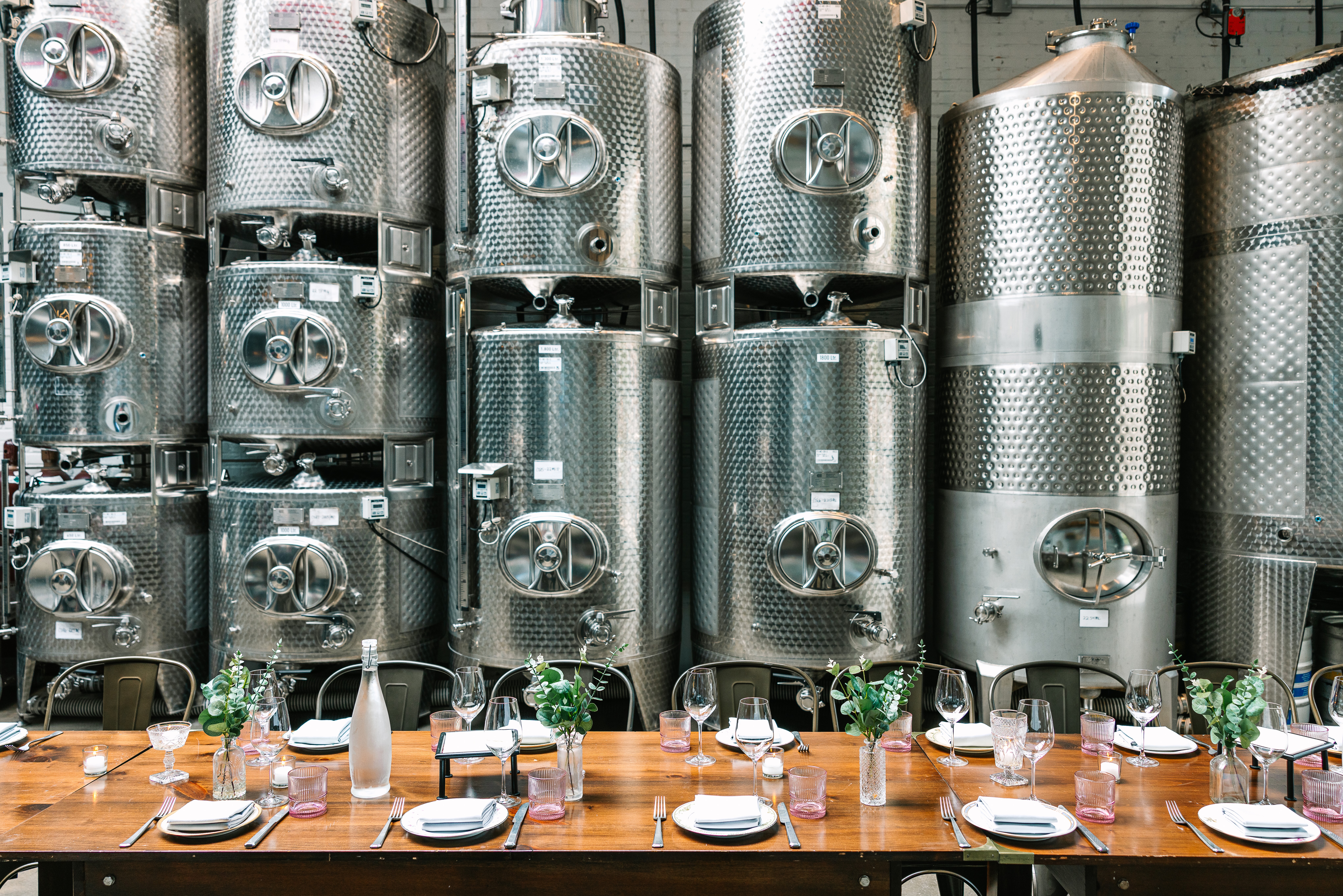 steel wine tanks in the background, in front of them is a long table set for a family style meal