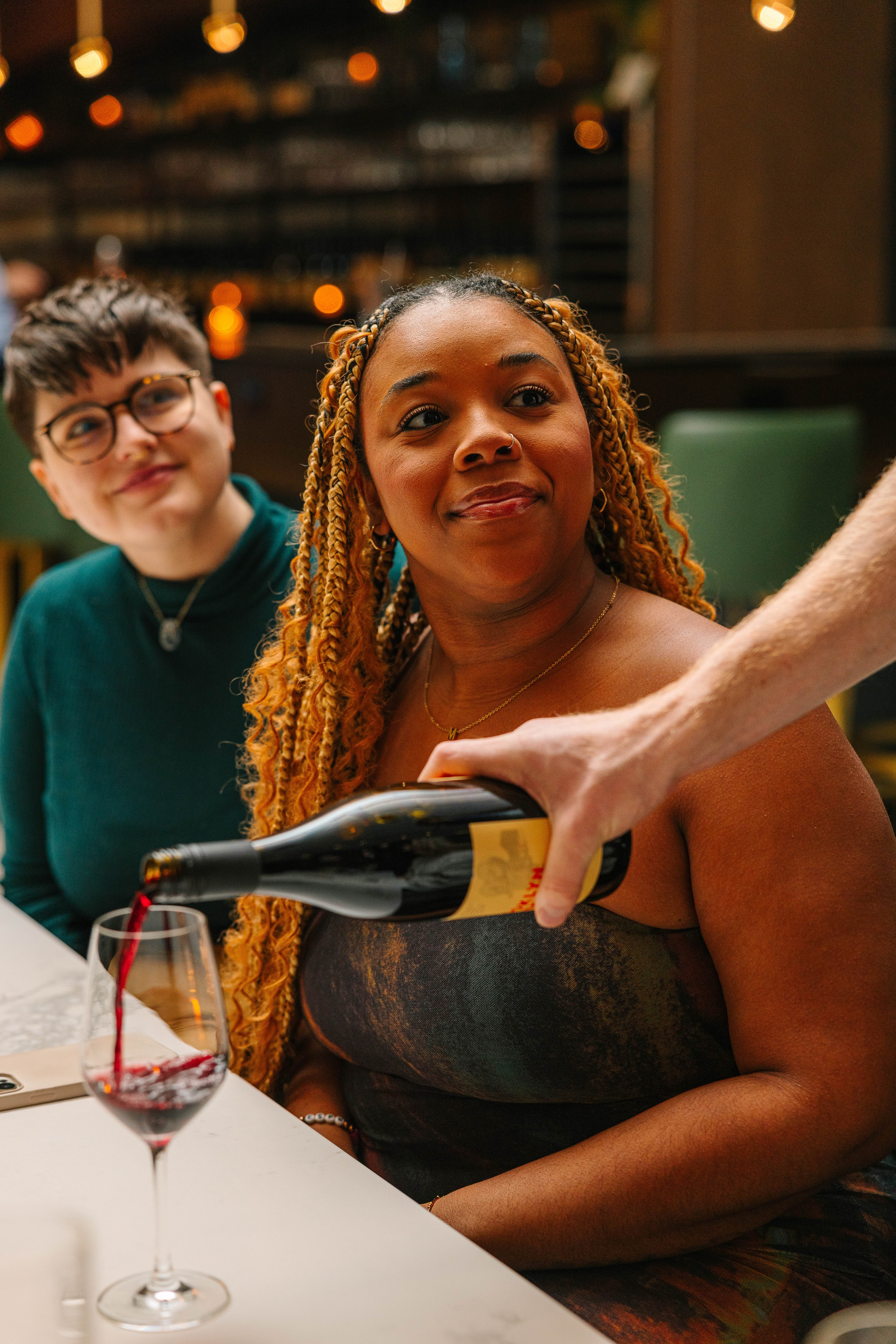 two guests watch as a server pours a glass of red wine