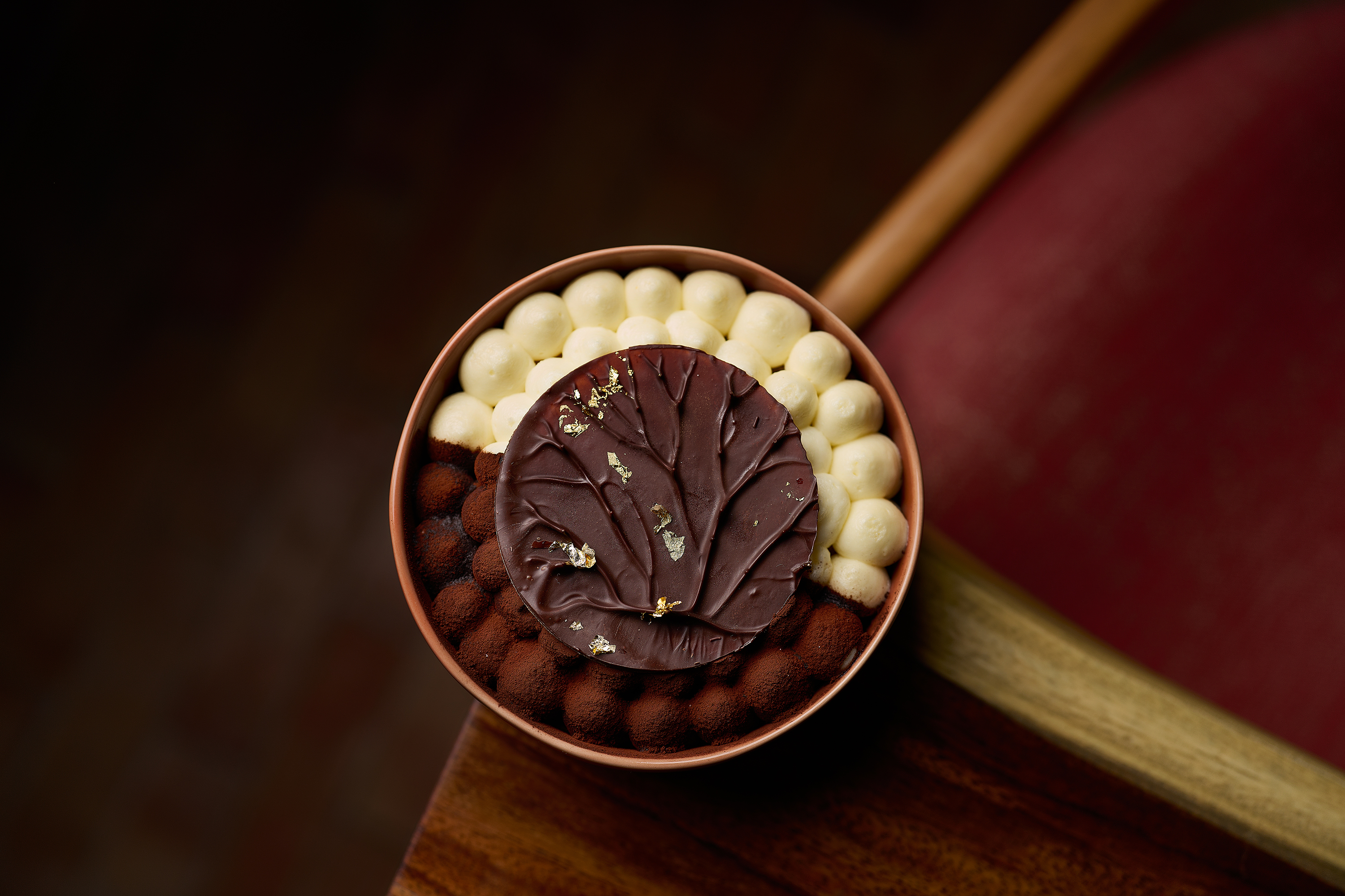 A round tiramisu served in a bowl, finished with a chocolate leaf and gold flakes