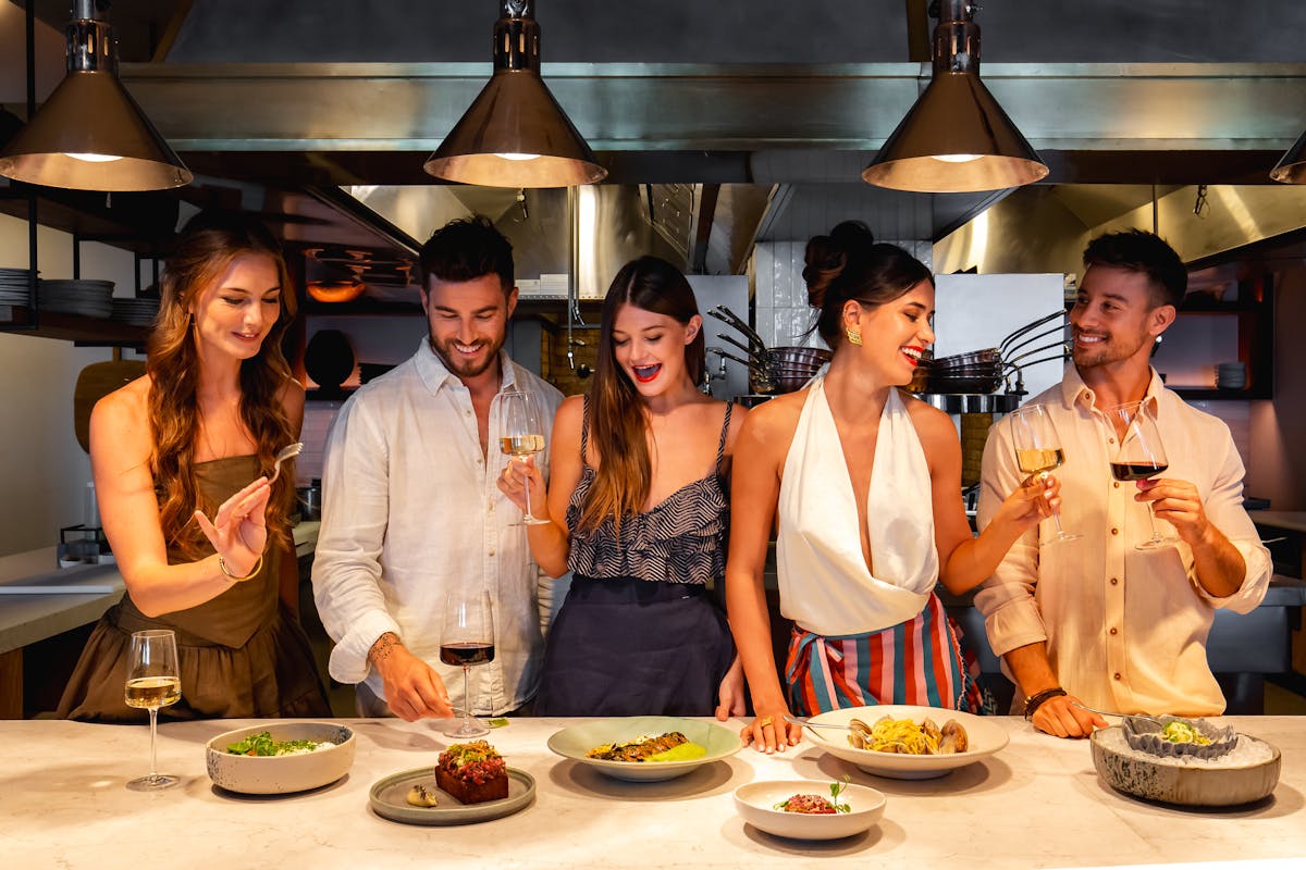 group of friends enjoying a lively dinner at Palmerio restaurant, toasting with wine and sharing Mediterranean-inspired dishes at the chef’s counter in Four Seasons Cabo San Lucas