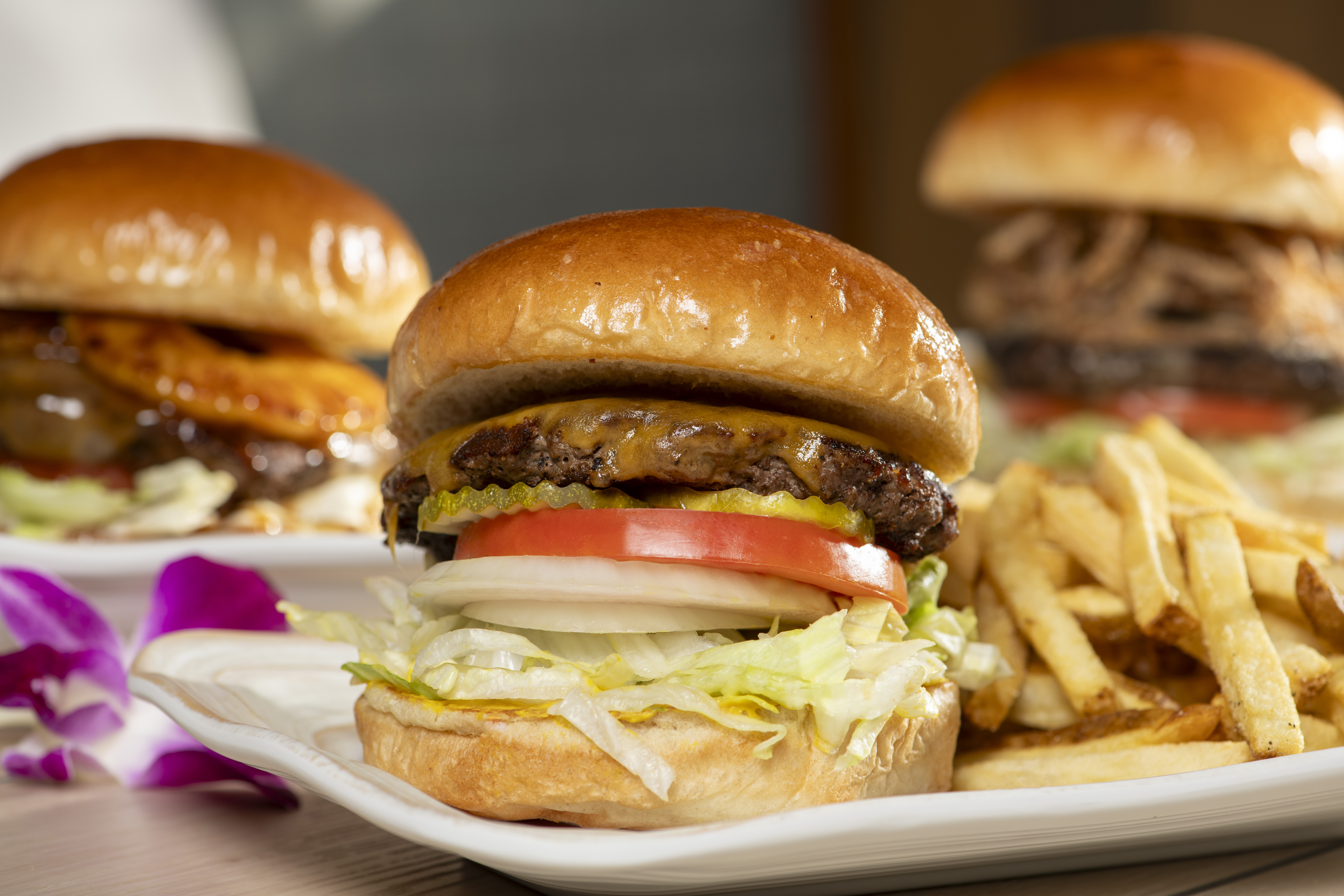 a close up of a cheeseburger on a plate with fries.  Two other burgers are in the background and are blurred out.