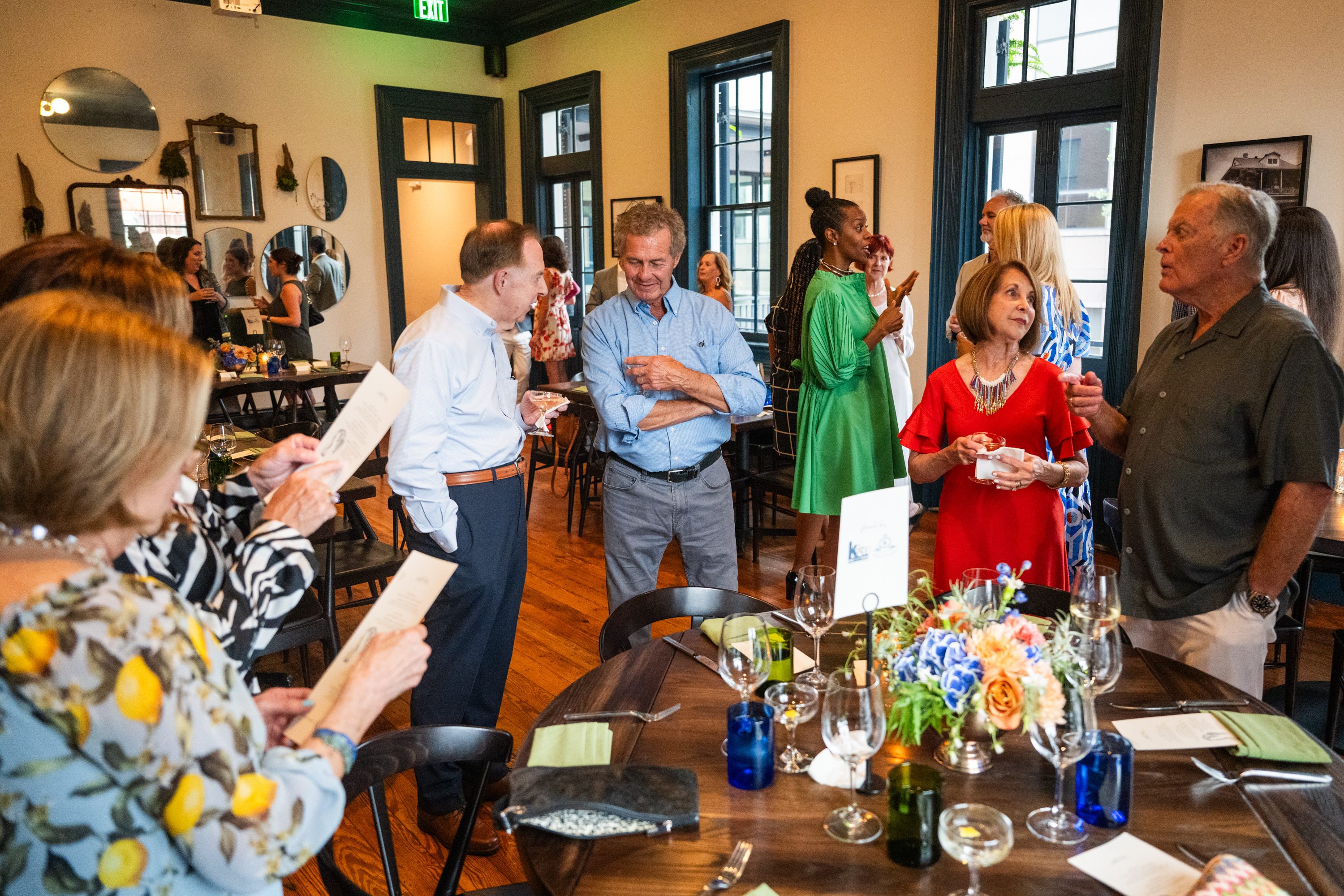 A group of people standing and socializing around a dinner table with wine glasses and flowers.