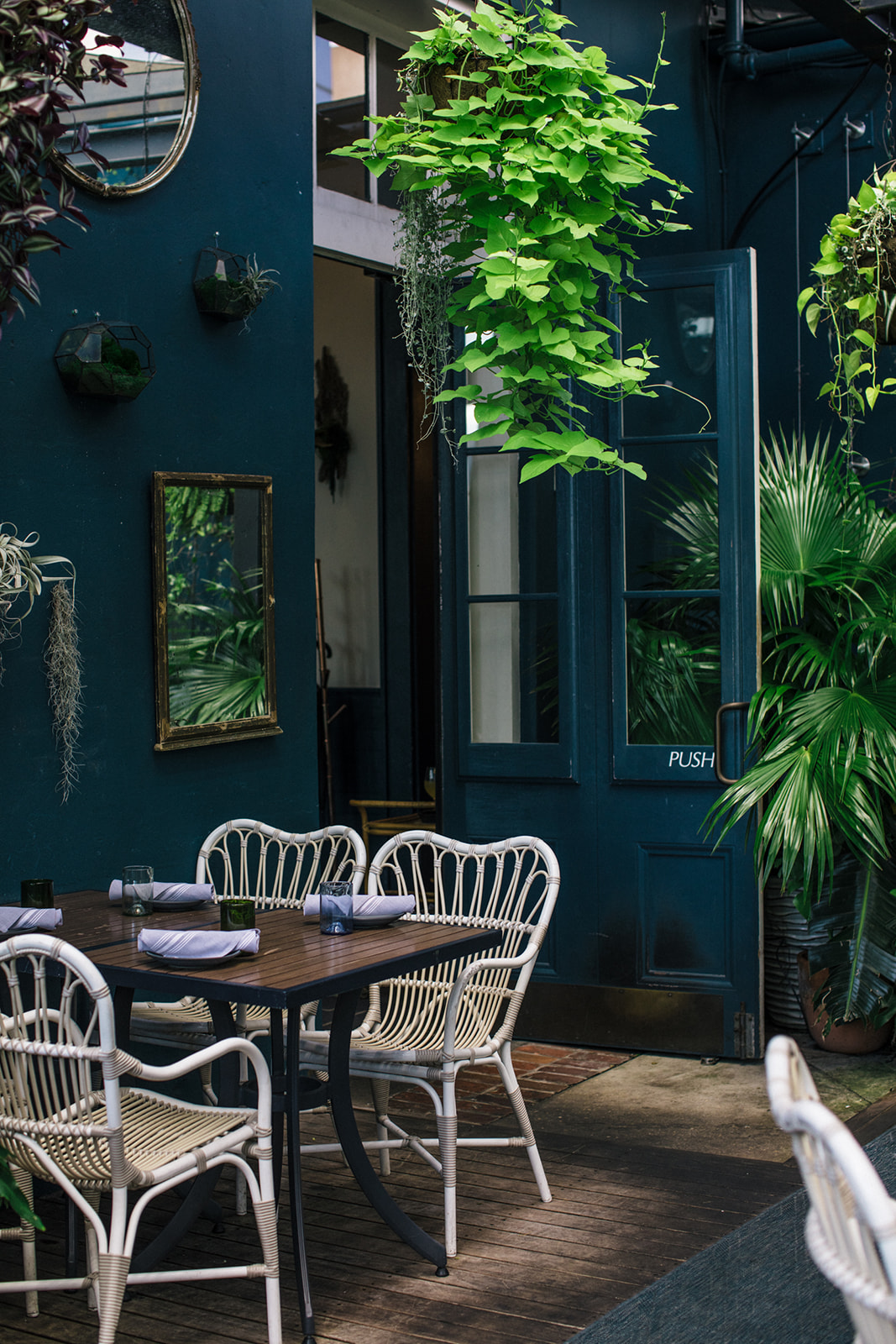 A cozy outdoor dining area with a dark blue wall, white furniture, and lush green plants.