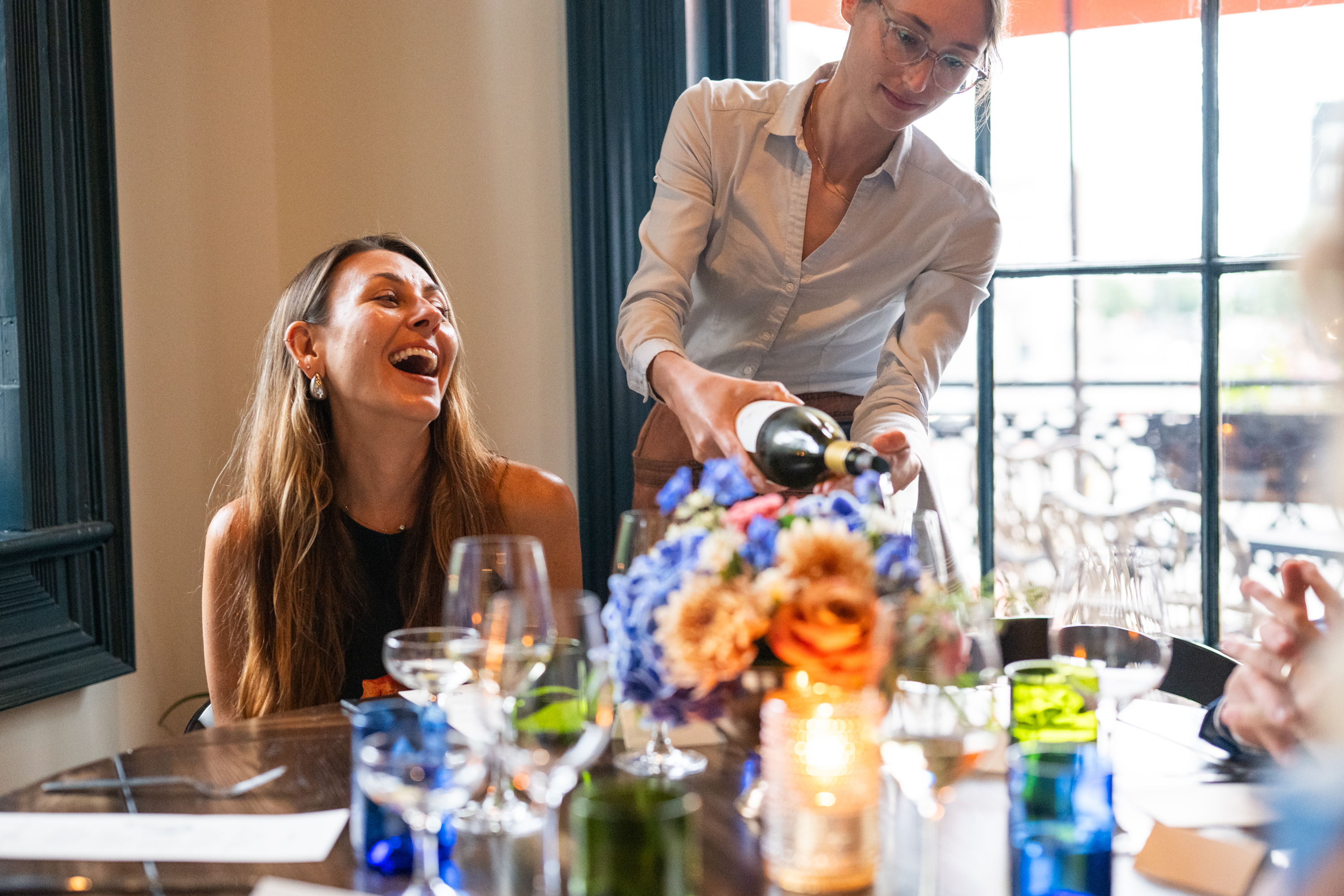 A woman laughs as another pours wine at a dining table.