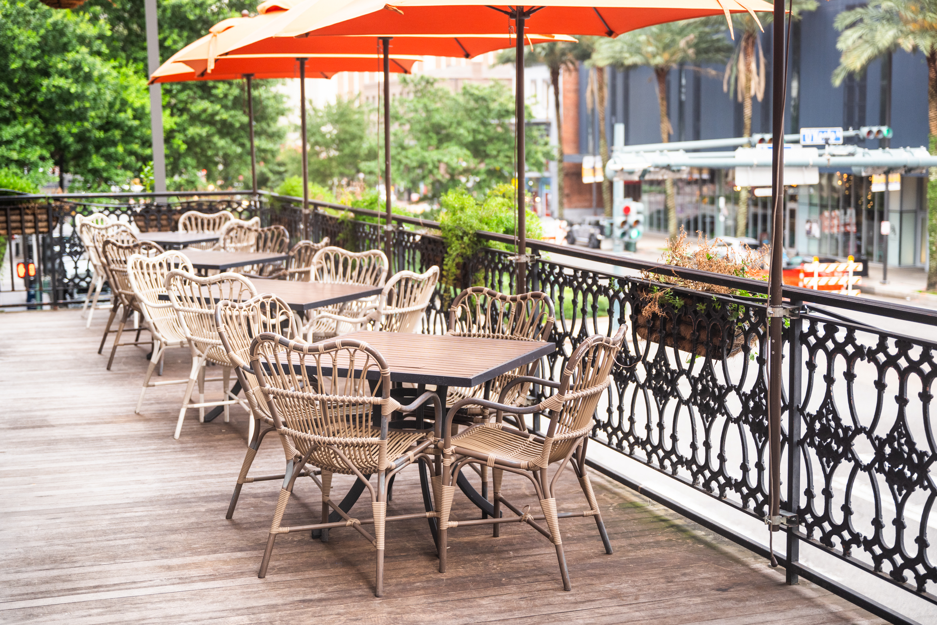 A wooden deck with outdoor dining tables and chairs, surrounded by a black railing and green trees.