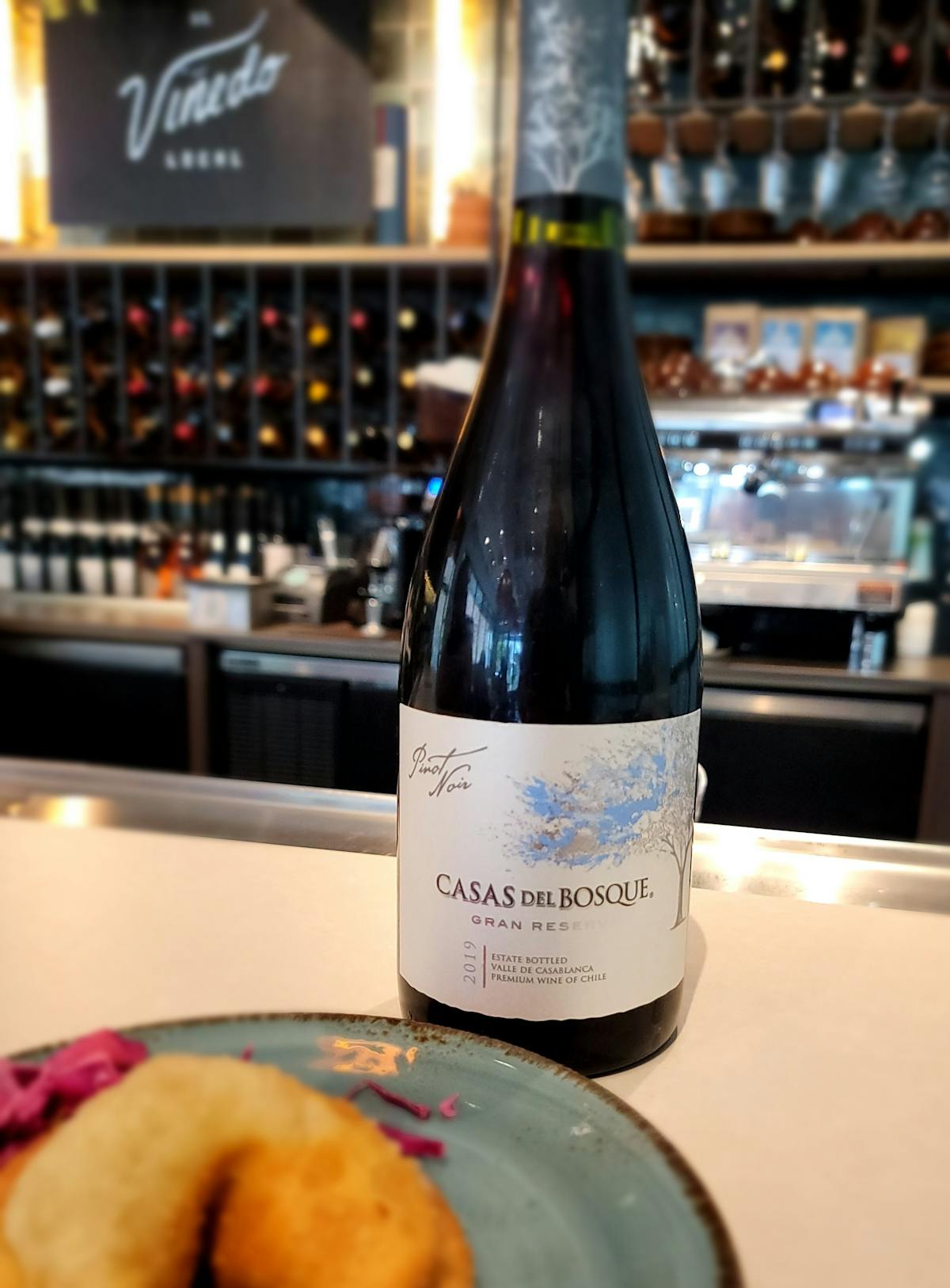 a bottle of Chilean pinot noir and a beef empanada at a bar with an espresso machine in the background