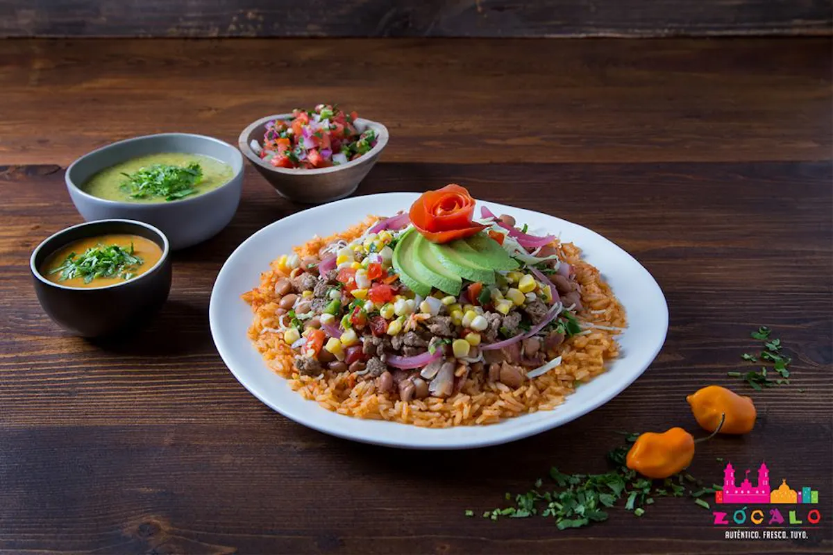 white plate layered with mexican rice, beans, corn, meat, topped with avocado sitting on a wood table with 3 bowls of various dips