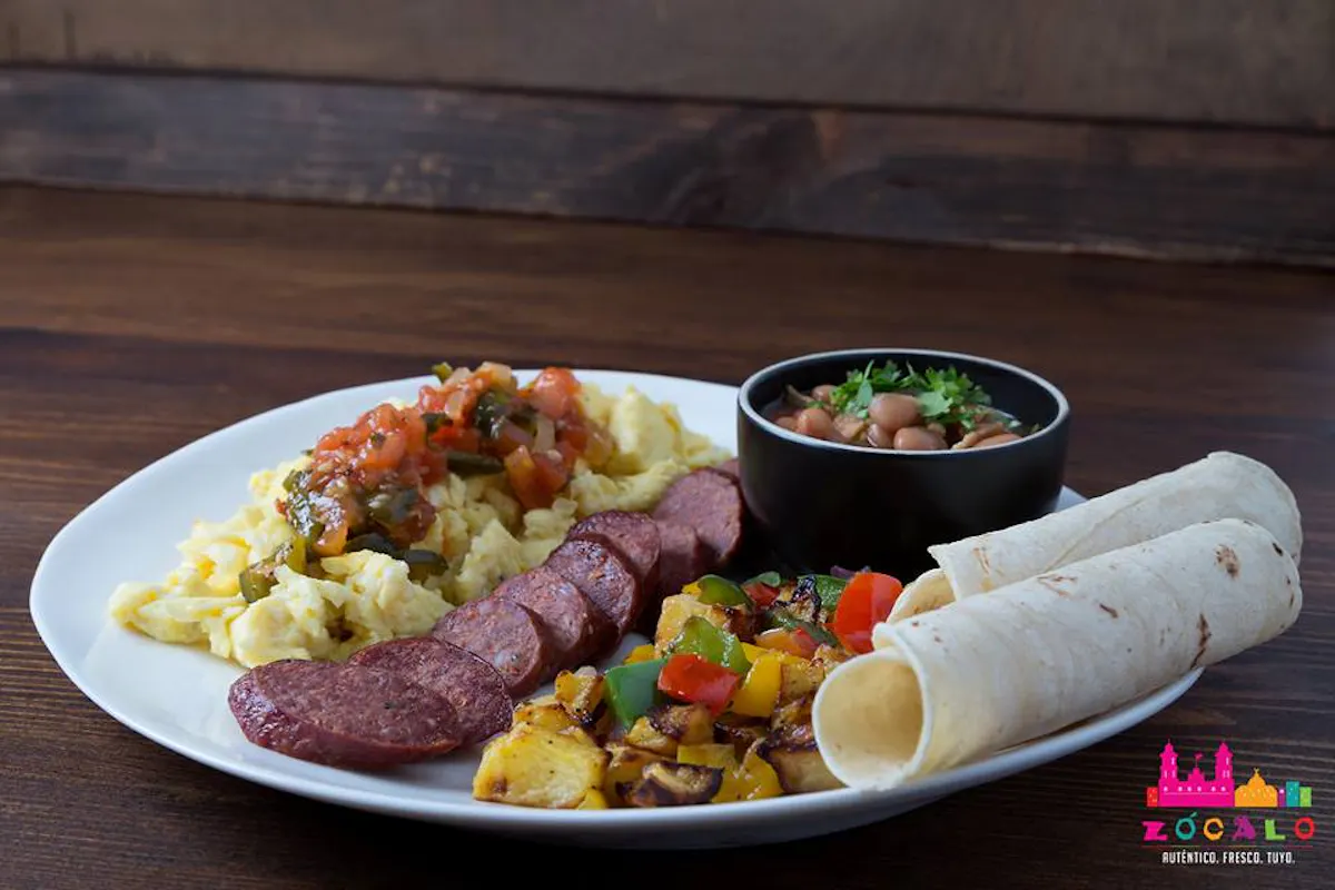 white plate of food consisting of corn tortillas, sliced meat, potatoes with peppers, small black bowl of beans.