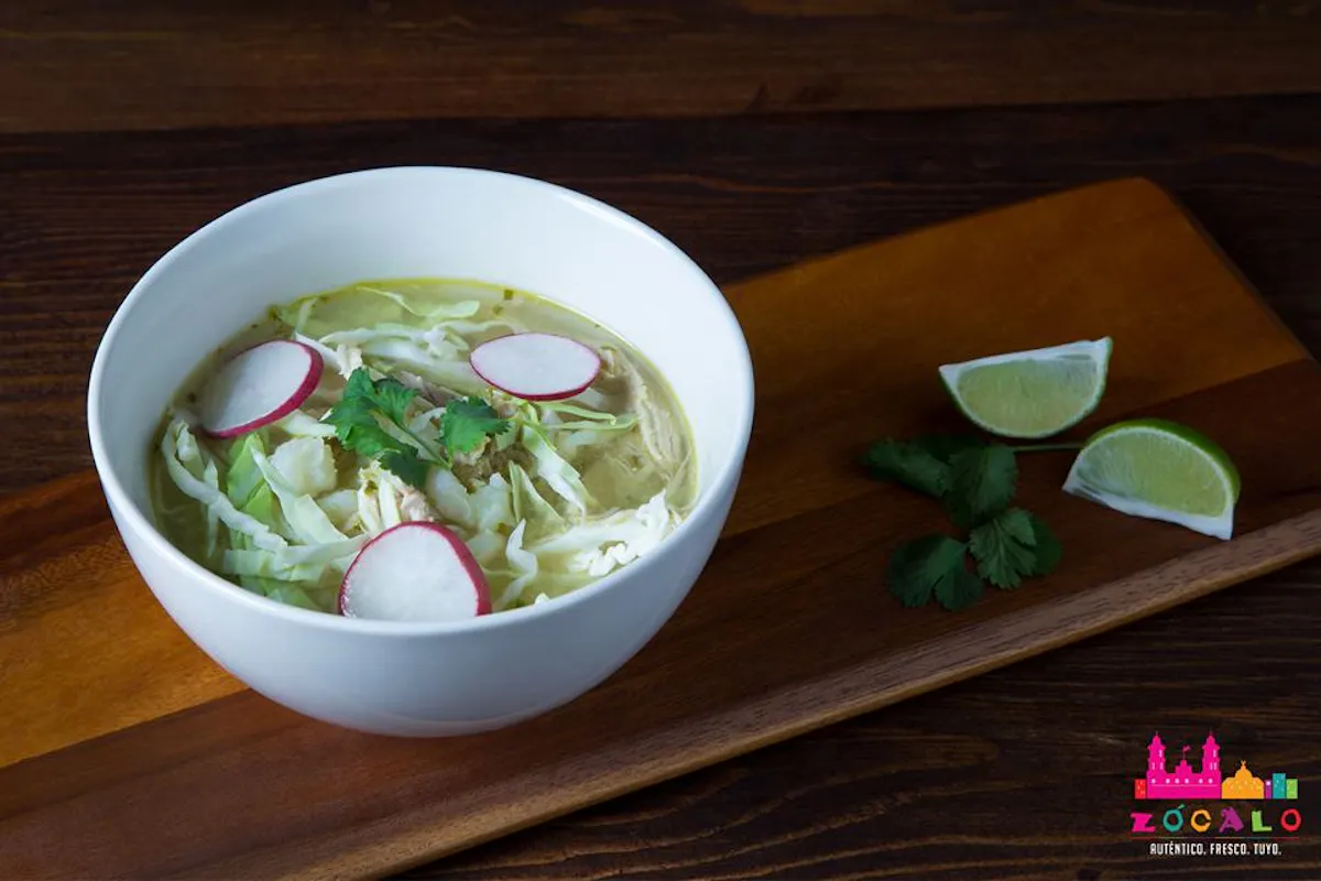 green broth soup with chicken topped with cilantro and radish. Bowl is placed on a wooden table with lime wedges in the background