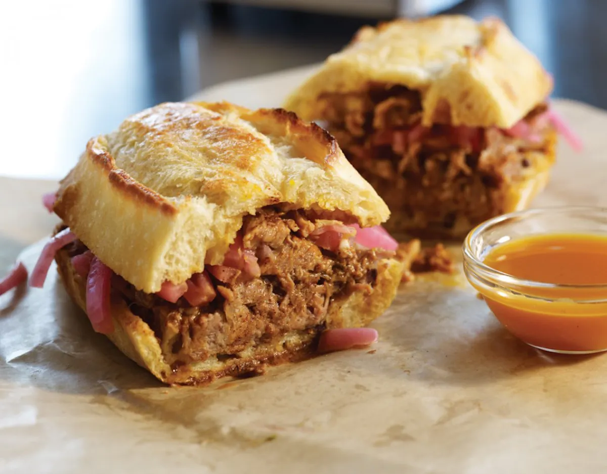 sandwich filled with meat and picled red onions, sitting on a cement table with a cup of sauce next to sandwich