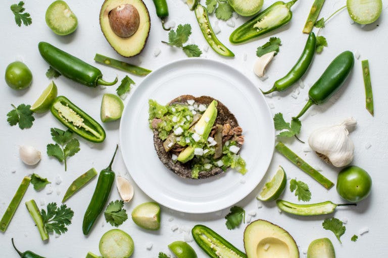 a blue tortilla taco on a plate surrounded by green ingredients such as avocado, chilly and leaves